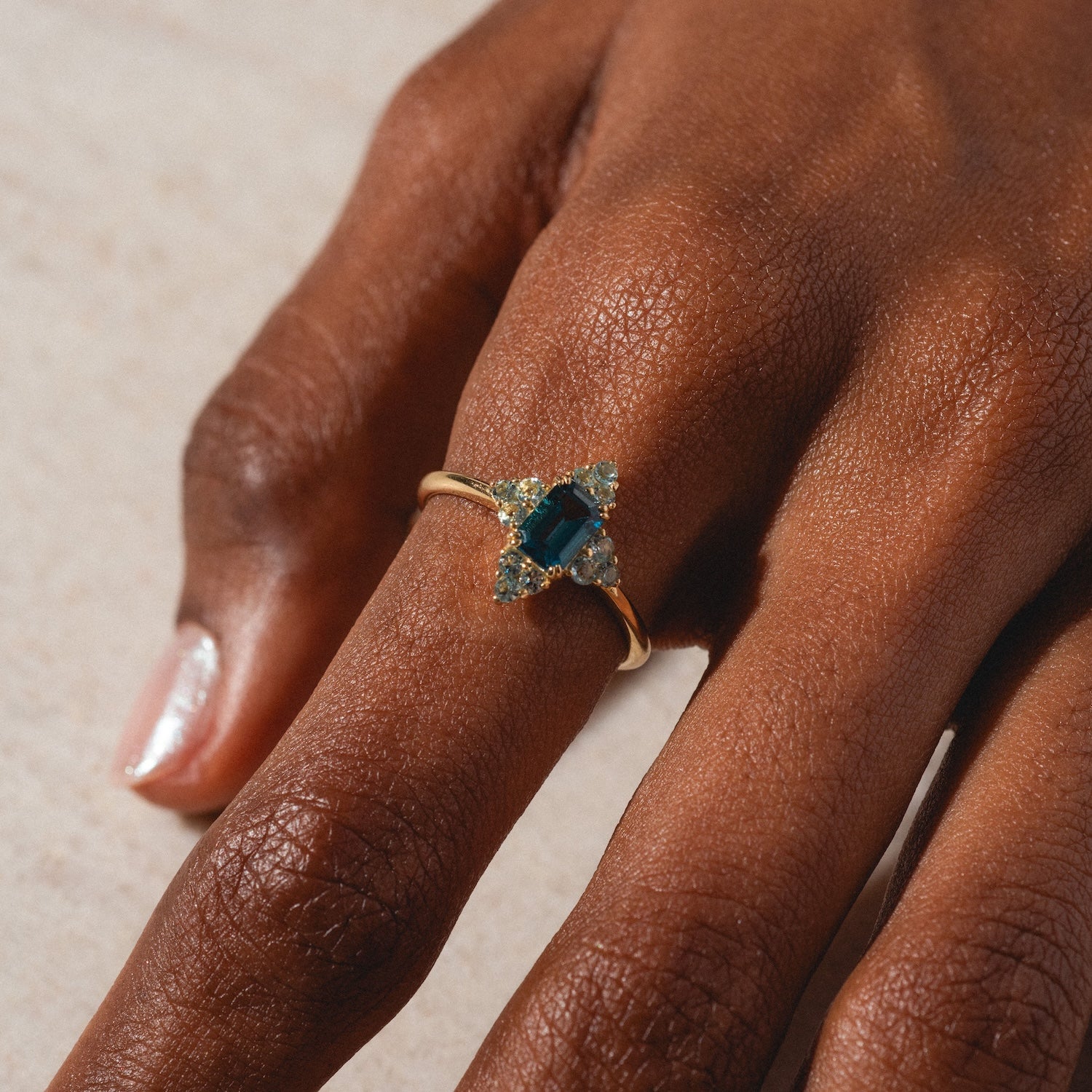 A close-up of a hand wearing the 10k Snow Queen Blue Topaz Cluster Ring, featuring a rectangular blue topaz center stone with two clusters of clear gemstones, shown against a light background.