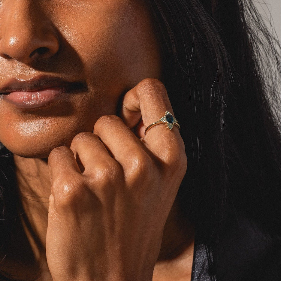 A woman with long dark hair, wearing a dark jacket, gazes at the camera with sunlight on her face and hand. She rests her chin on her hand, showcasing the 10k Snow Queen Blue Topaz Cluster Ring on her finger.