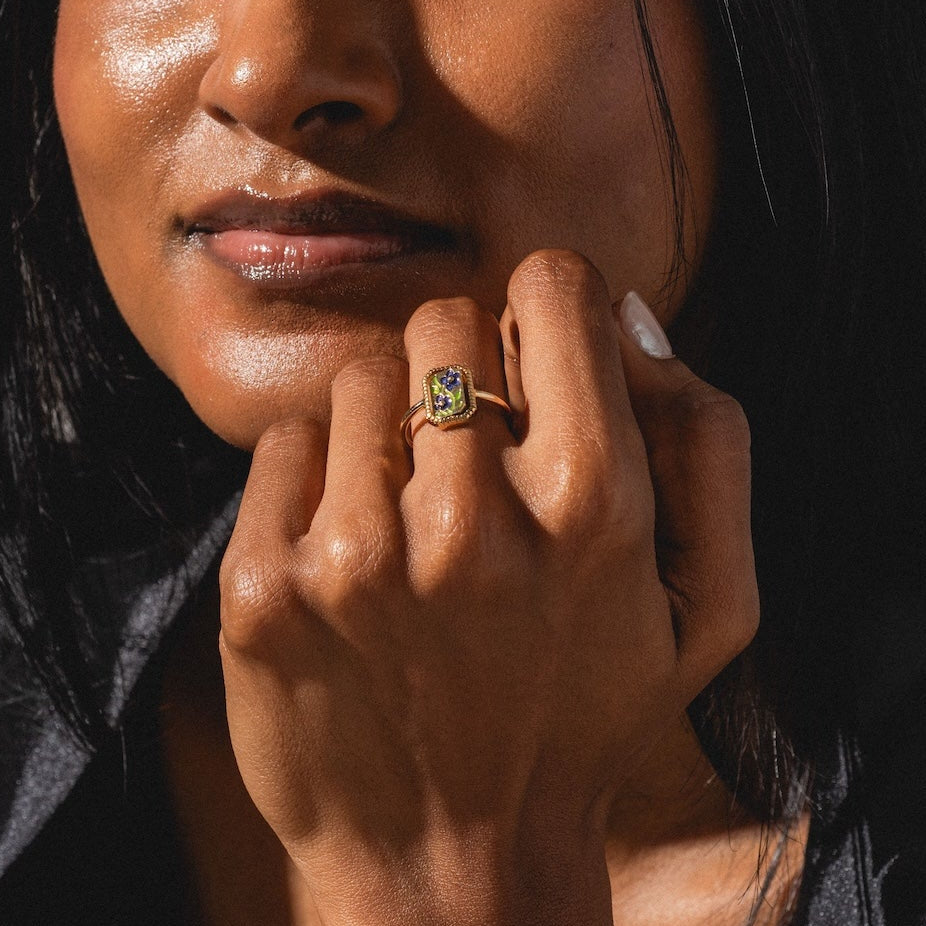A woman with long dark hair and radiant skin smiles gently, wearing the 10k Gallery Frames Enamel and Malachite Reversible Ring. Dressed in a silky collared shirt, sunlight illuminates her face as she rests her chin on her hand.