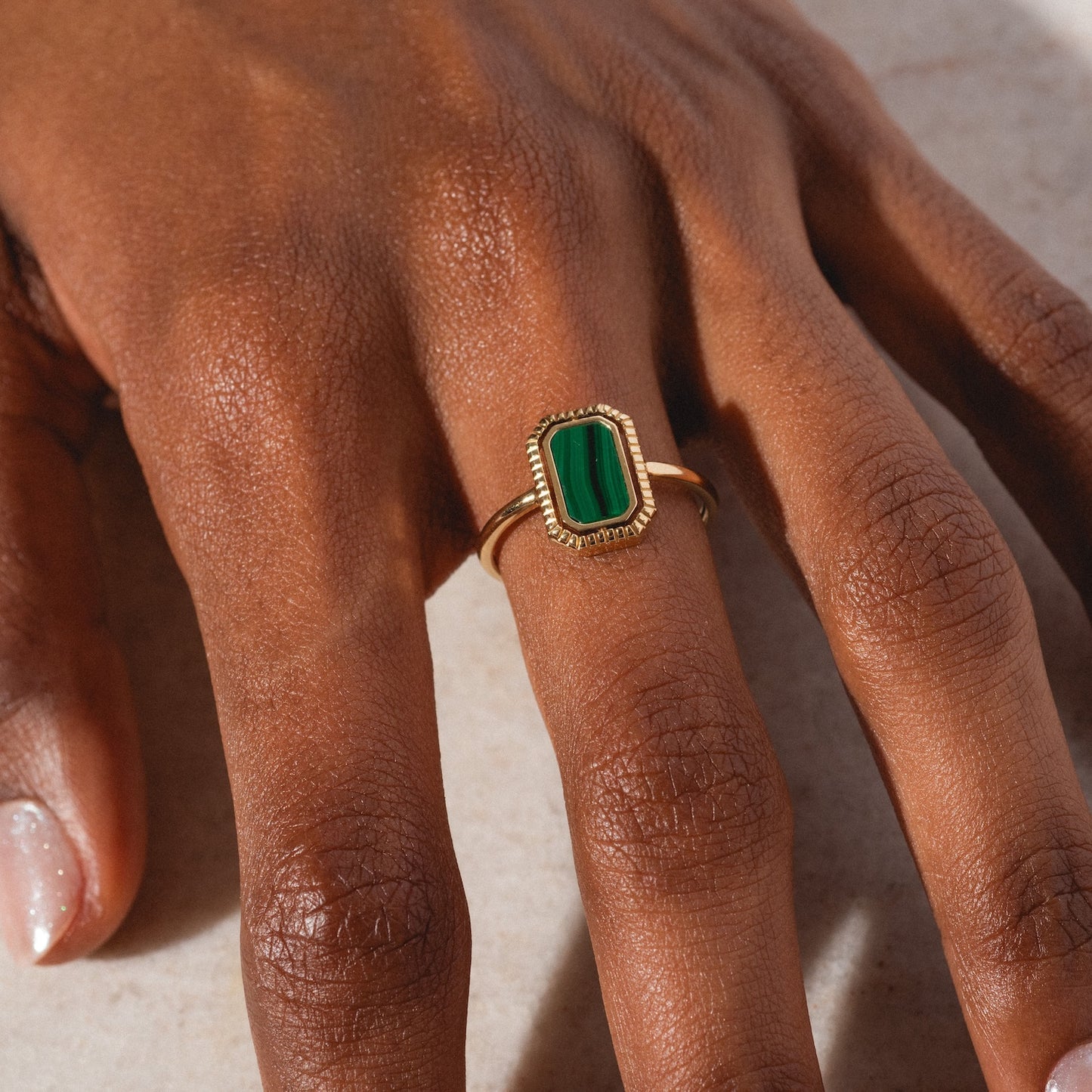 A hand with medium brown skin wears the 10k Gallery Frames Enamel and Malachite Reversible Ring, showcasing a rectangular green malachite stone with a textured gold border, resting on a light surface.