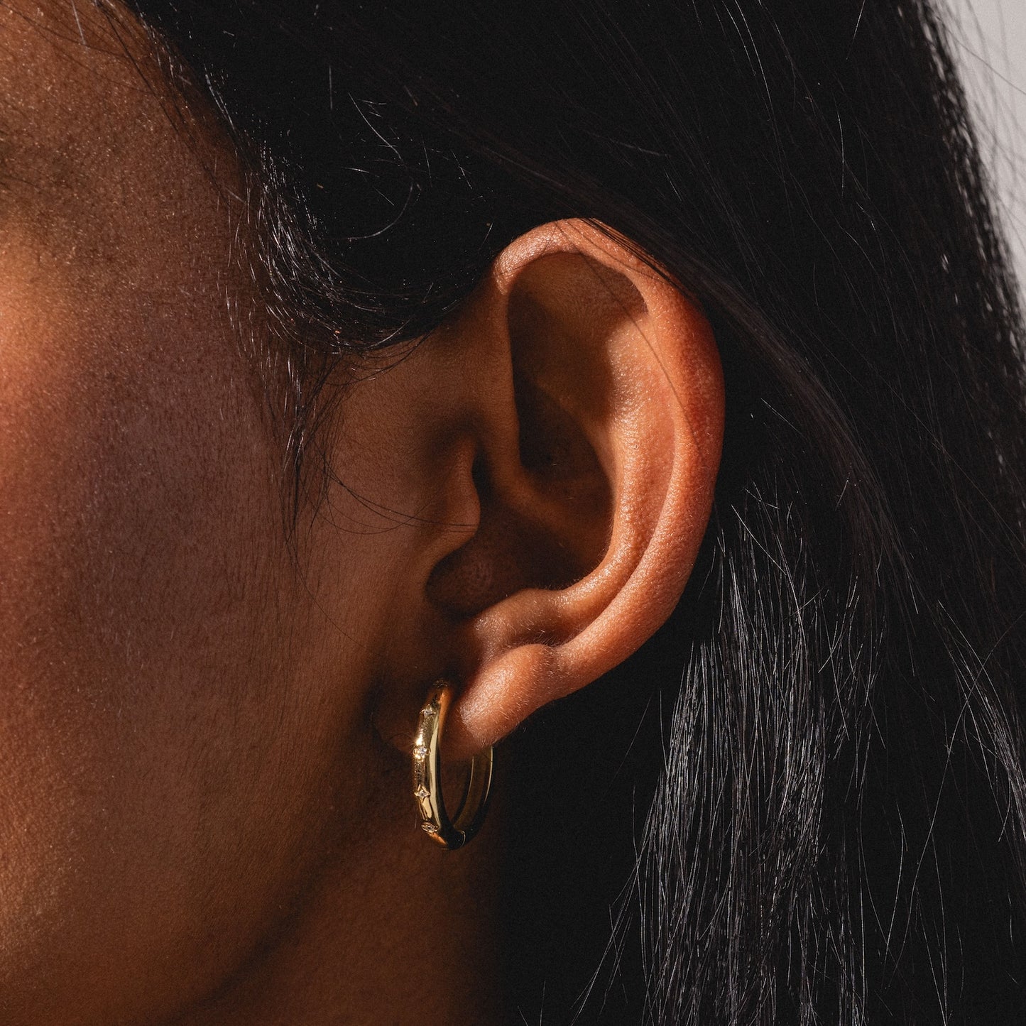 Close-up of a person’s ear with long, dark hair wearing a Bold Birthstone Earring Set. The lighting highlights the earring and ear against a softly blurred background.