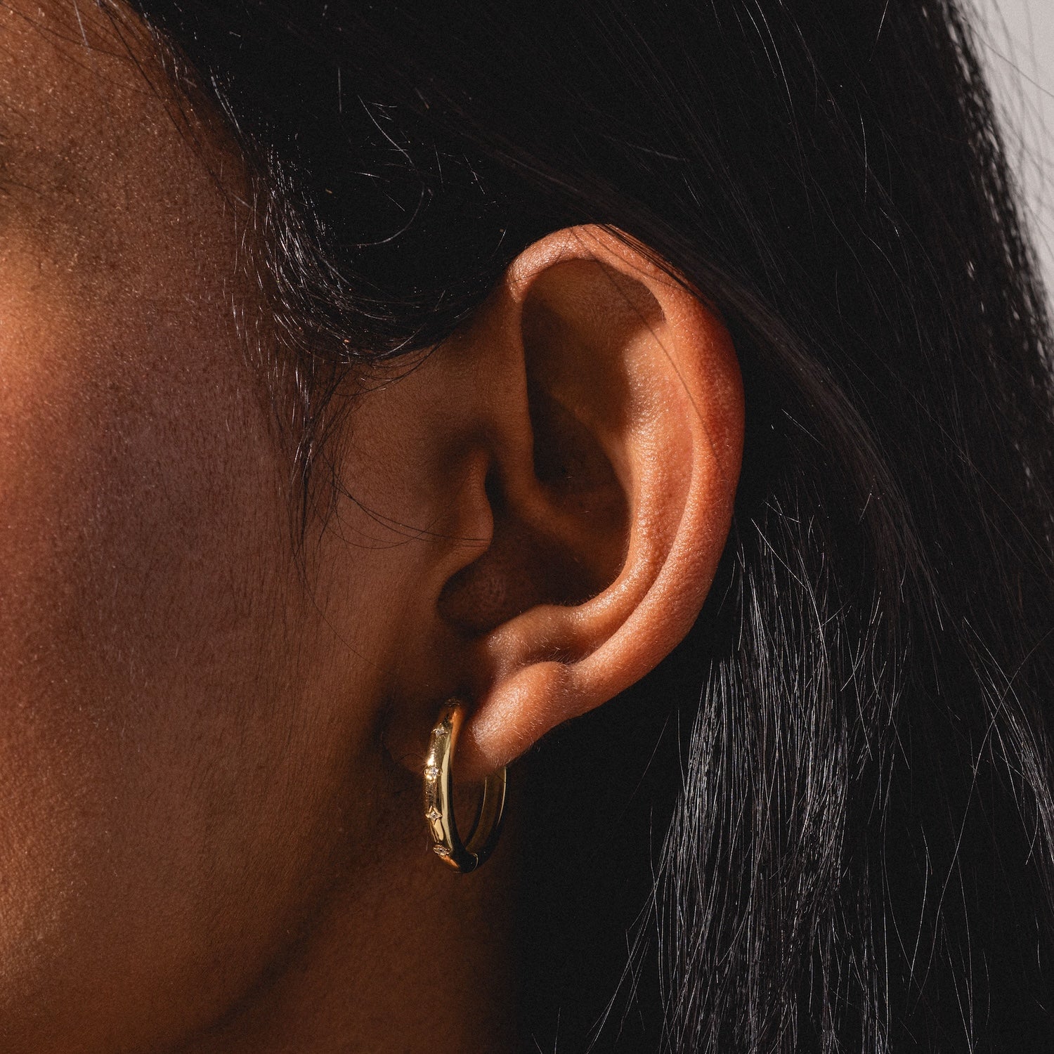 Close-up of a person’s ear with long, dark hair wearing a Bold Birthstone Earring Set. The lighting highlights the earring and ear against a softly blurred background.