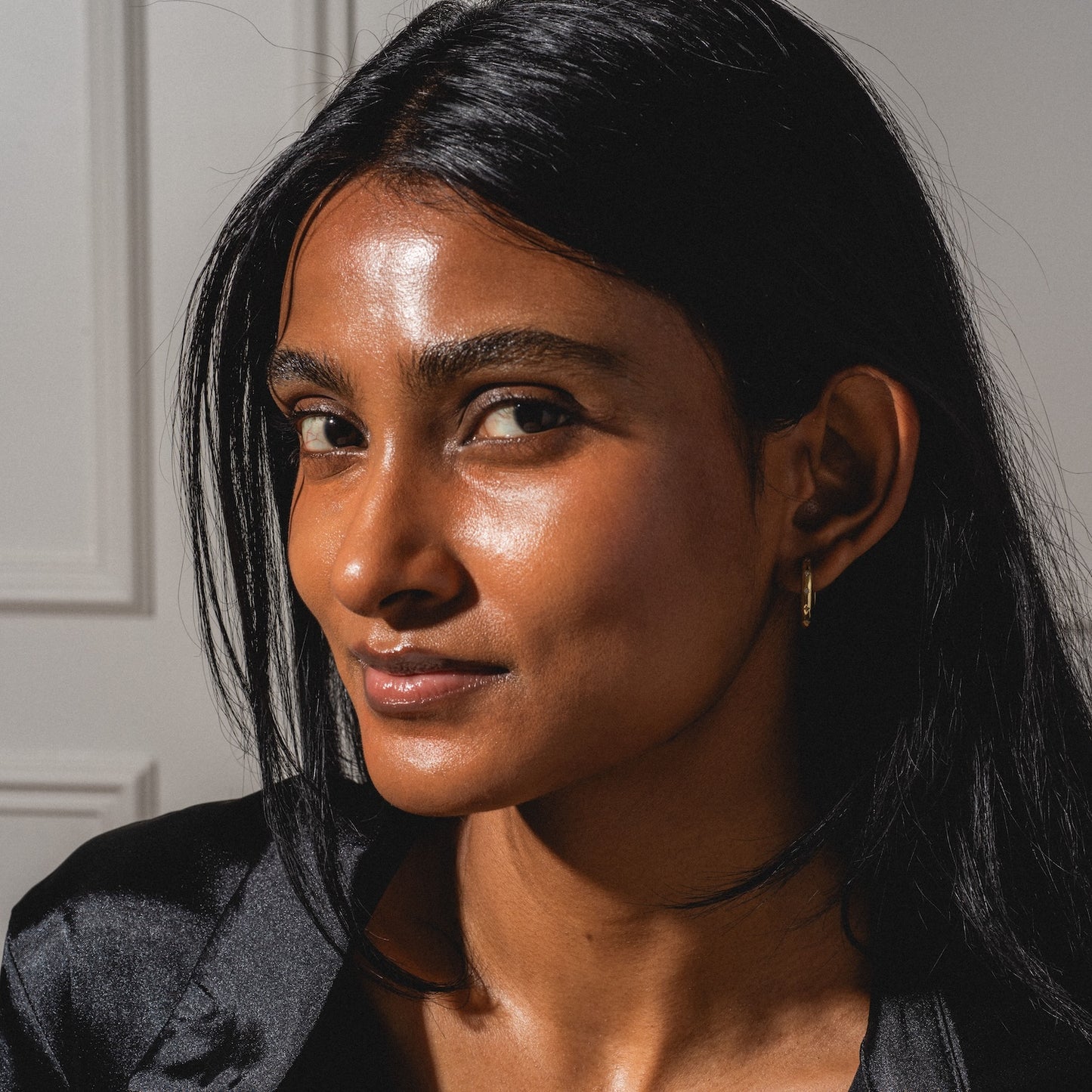 A woman with long black hair and glowing brown skin looks confidently at the camera, wearing a black top and the Bold Birthstone Earring Set, with sunlight highlighting her features against a neutral background.