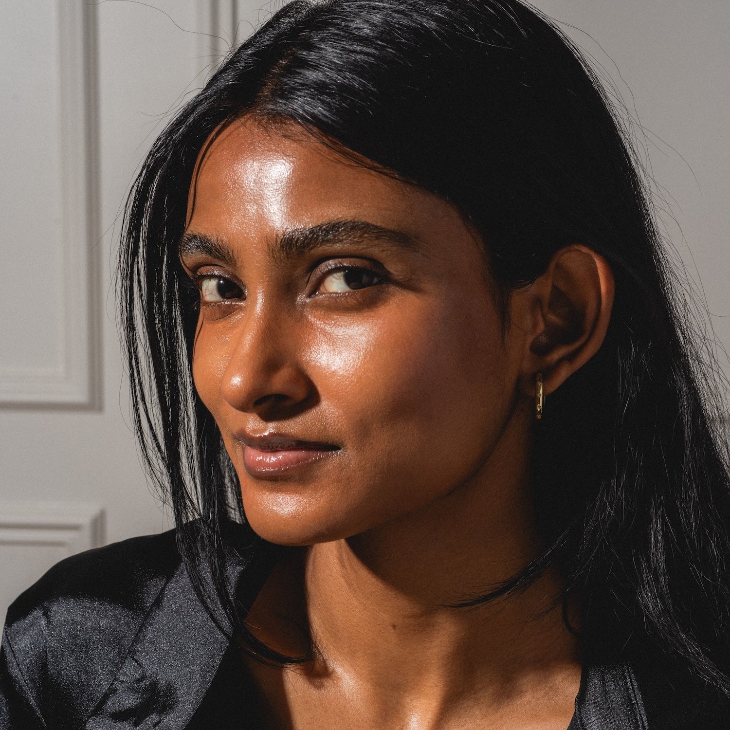 A woman with long black hair and glowing brown skin looks confidently at the camera, wearing a black top and the Bold Birthstone Earring Set, with sunlight highlighting her features against a neutral background.