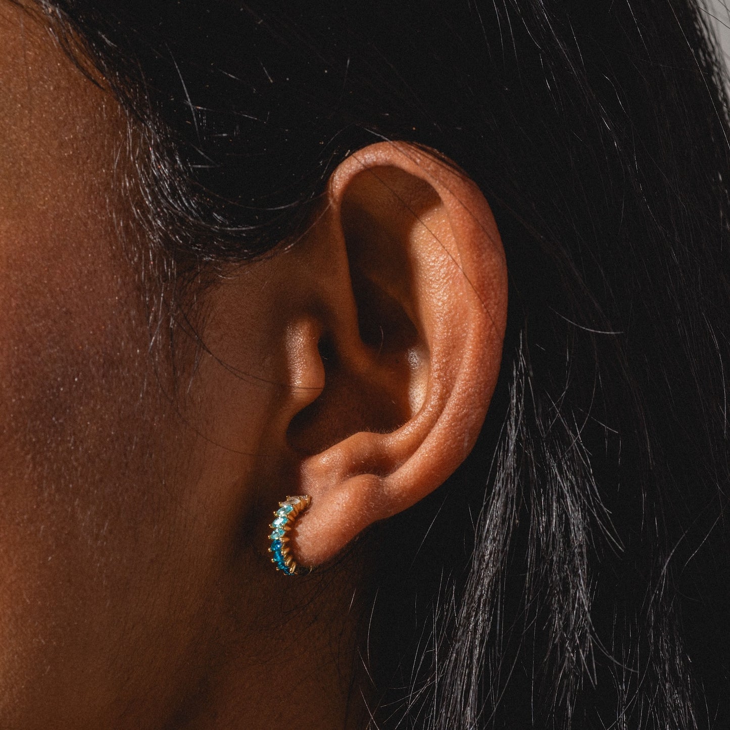 A close-up of a person’s ear with long dark hair wearing a small gold hoop from the Bold Birthstone Earring Set, adorned with turquoise-colored stones. Warm sunlight highlights both the skin and the elegant jewelry.