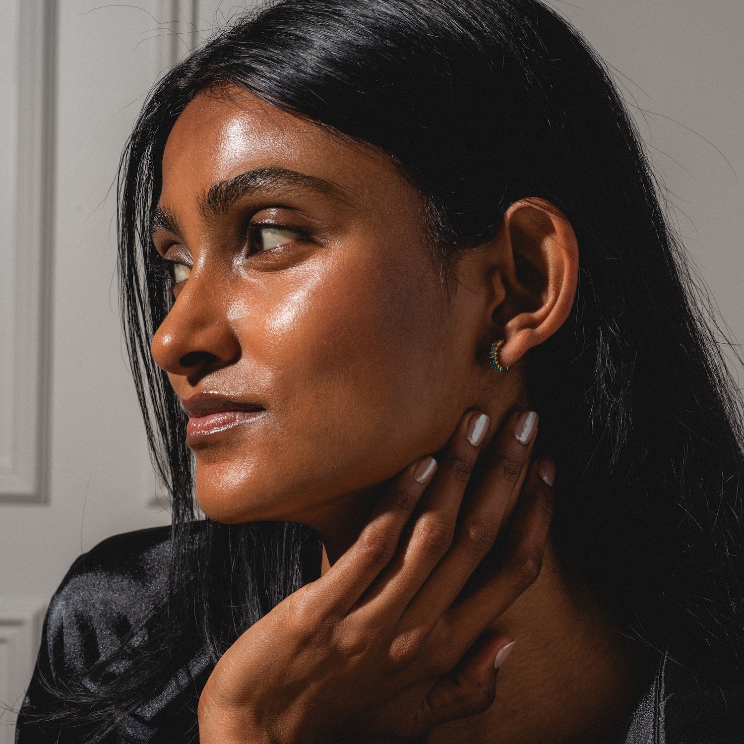 A woman with long black hair and glowing skin looks to the side, wearing the Bold Birthstone Earring Set. She gently touches her neck, polished nails visible, while soft light highlights her features against a neutral backdrop.