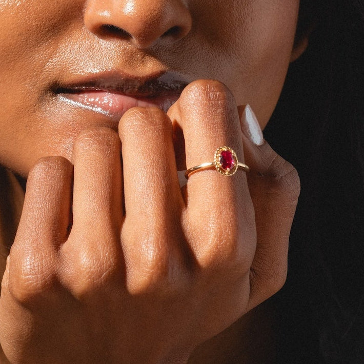 A woman with long dark hair gazes thoughtfully at the camera, resting her chin on her hand while wearing the 10k Lucky In Red Ruby Halo Ring; sunlight highlights both her face and the sparkling red gemstone on her finger.