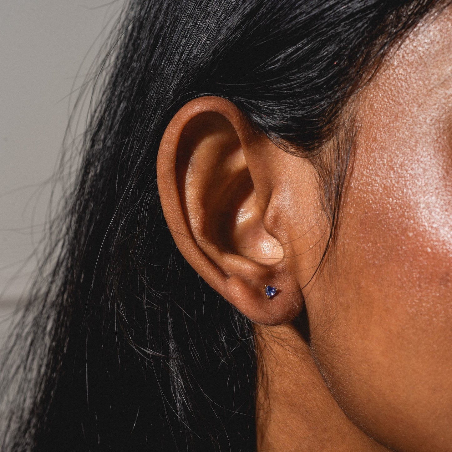 A close-up shows a person with smooth, medium-brown skin and straight, dark hair wearing a small, dark stud from the Bold Birthstone Earring Set in their earlobe.