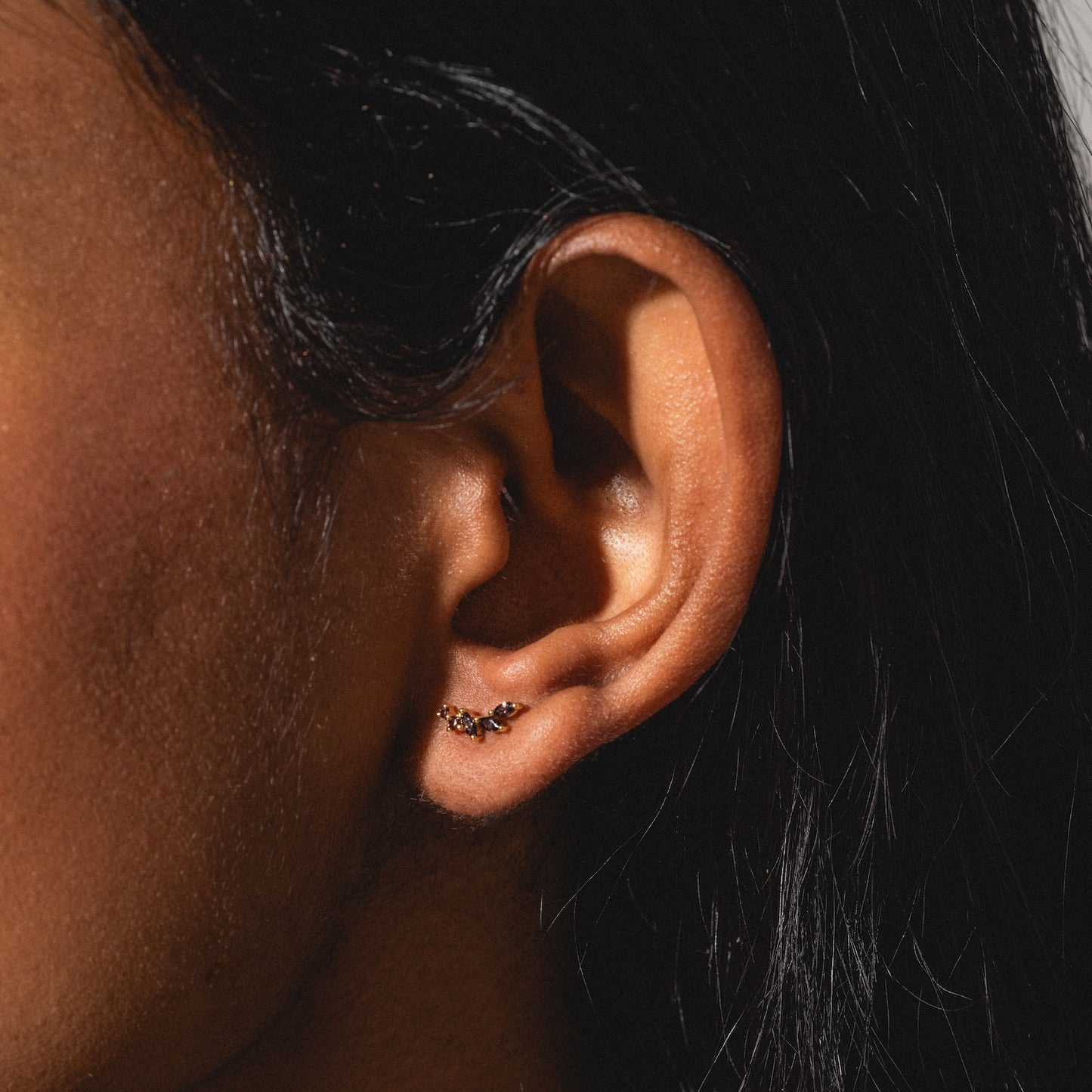 Close-up of an ear with straight dark hair, wearing a small gold Classic Birthstone Earring adorned with tiny sparkling stones on the earlobe, set against a neutral background.
