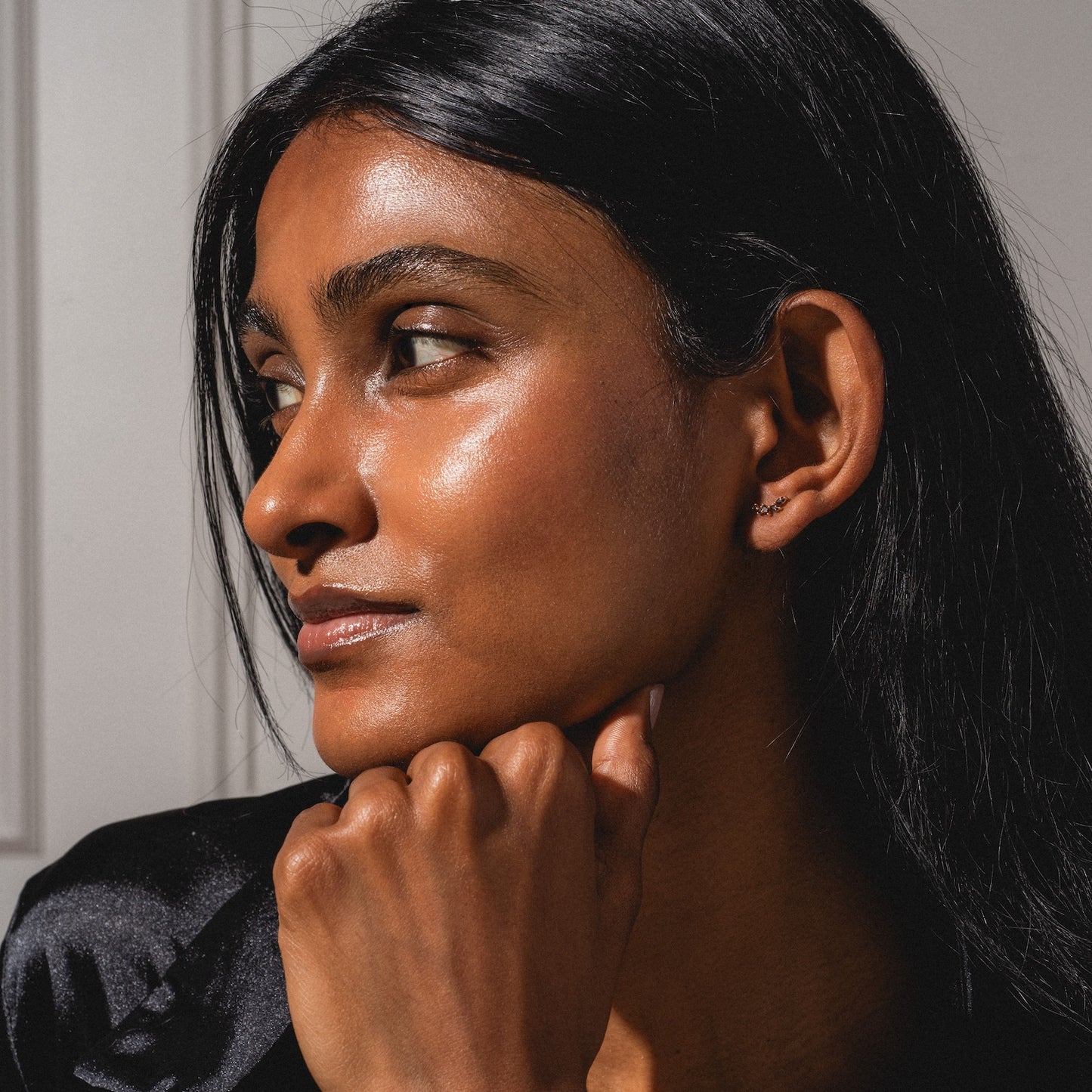 A woman with long dark hair and radiant skin gazes thoughtfully to the side, her chin resting on her hand. She wears a black top and a Classic Birthstone Earring Set, sunlight softly illuminating her face.