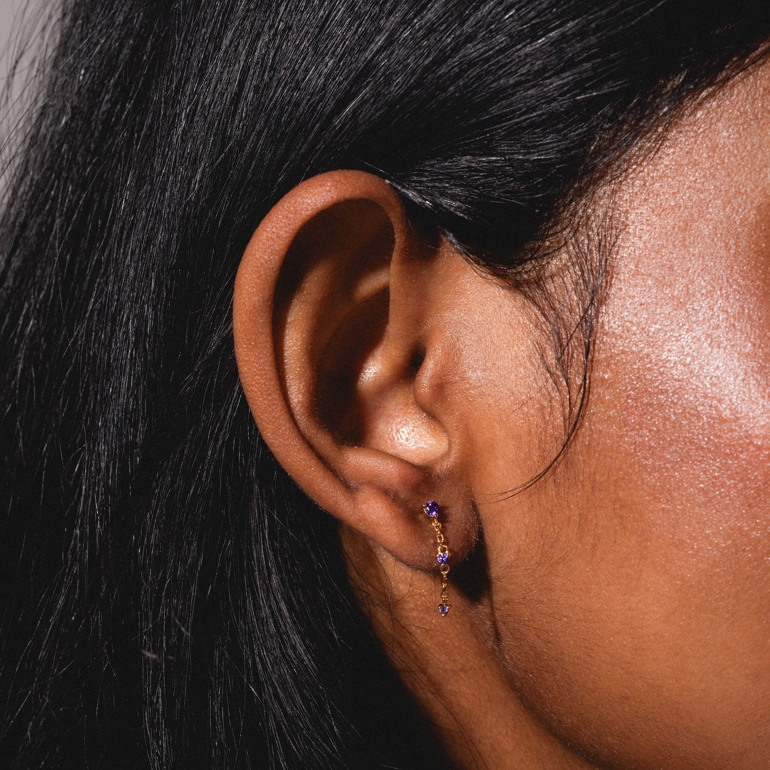 A close-up of a person with long, straight black hair wearing the Classic Birthstone Earring Set—delicate gold earrings with small purple gemstones. Their skin is smooth and warm brown.