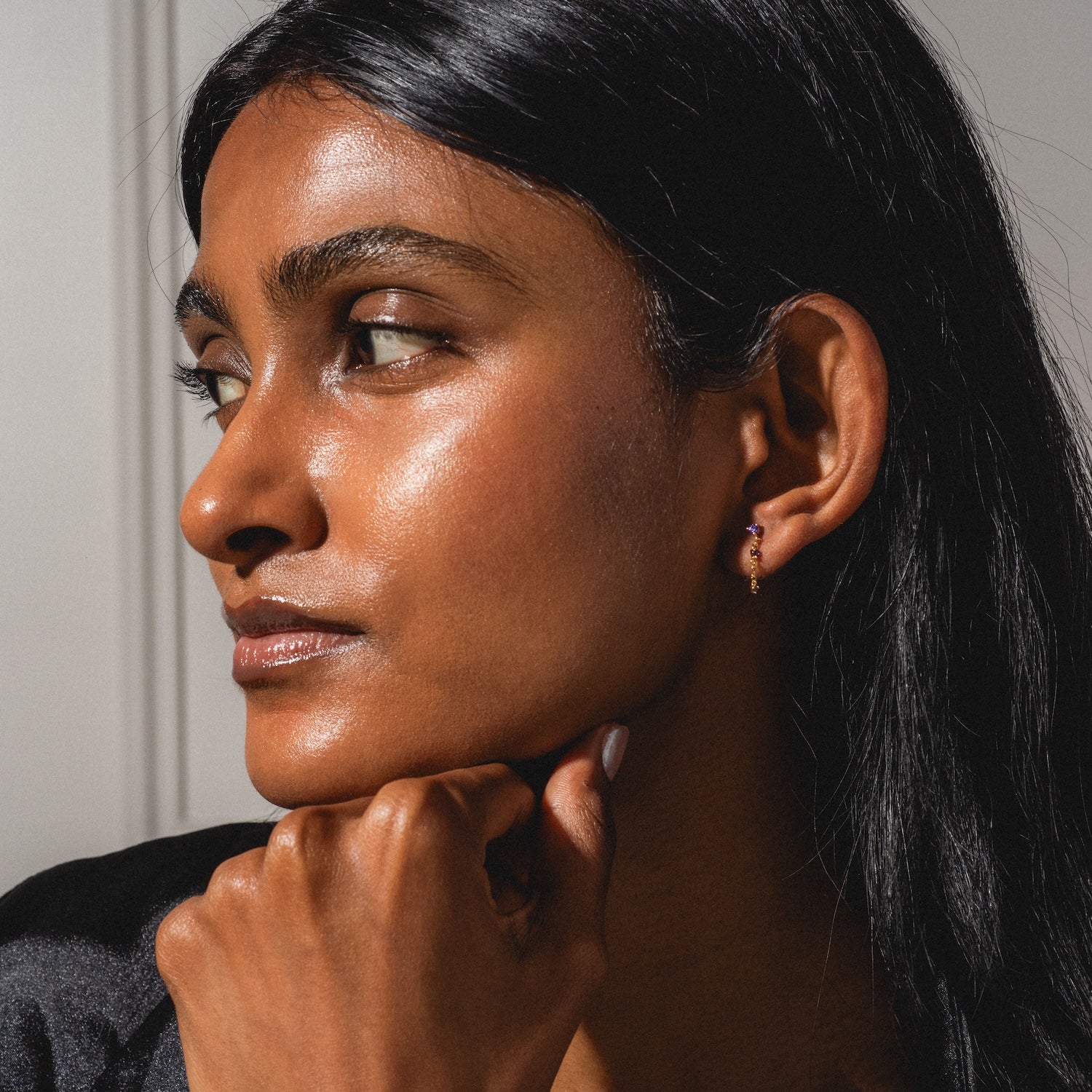 A woman with long dark hair and glowing skin gazes thoughtfully to the side, resting her chin on her hand. Sunlight highlights her face as she wears a Classic Birthstone Earring Set.