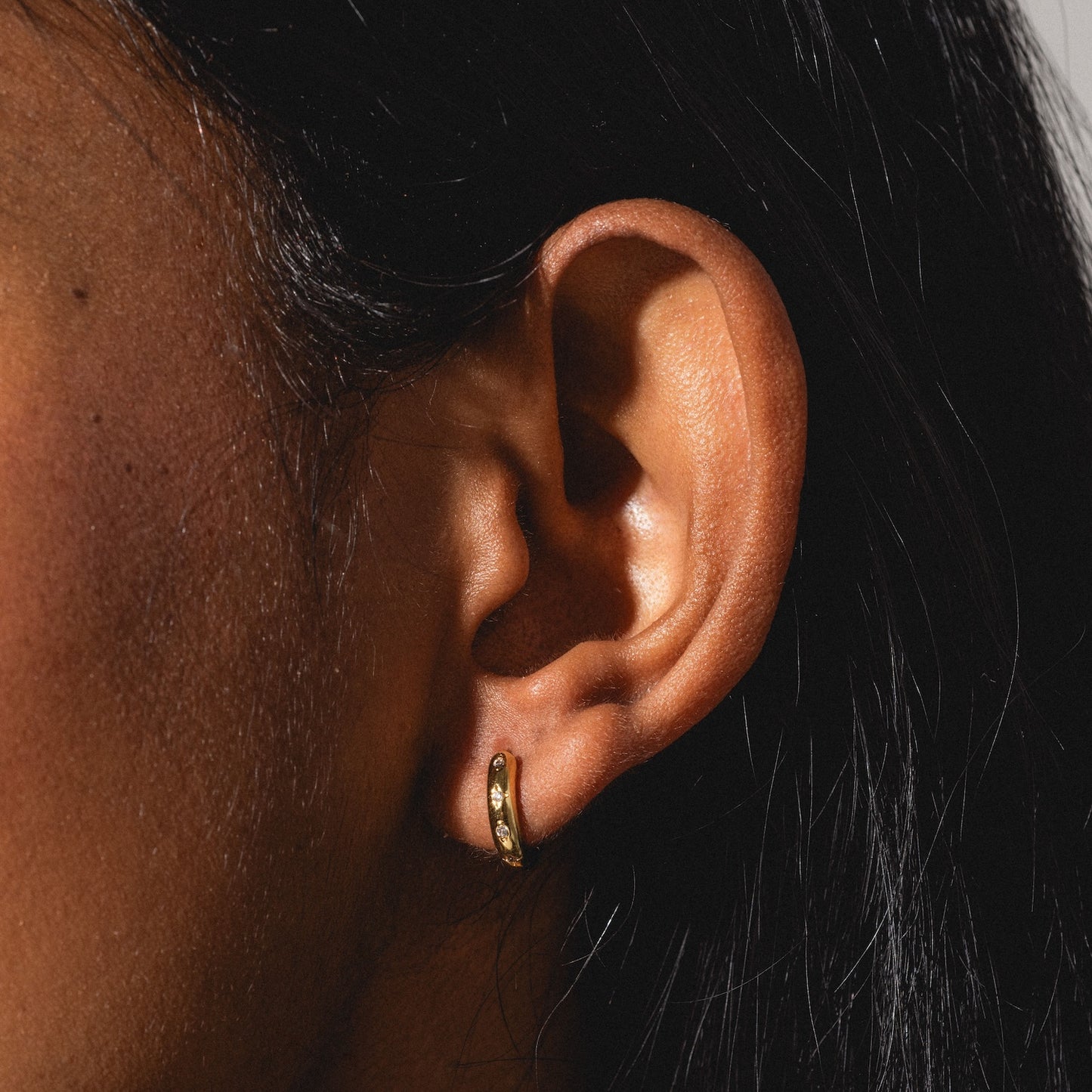 Close-up of a person with dark hair wearing a Classic Birthstone Earring, a small gold hoop set with sparkling stones. The lighting accentuates the earring’s detail and the ear’s texture.