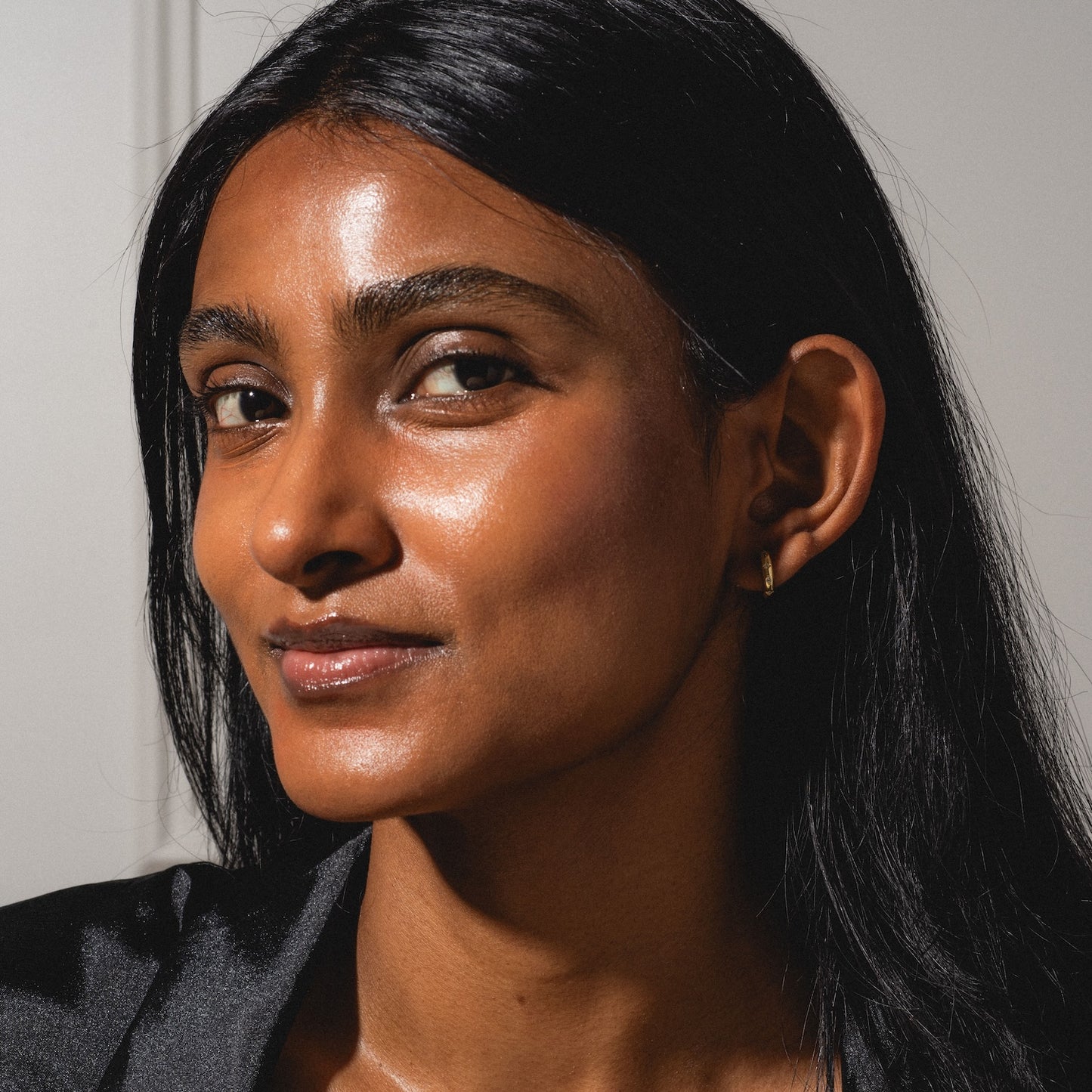 A woman with long, straight black hair and radiant skin looks confidently at the camera, wearing a black top and the Classic Birthstone Earring Set, softly illuminated against a neutral background.