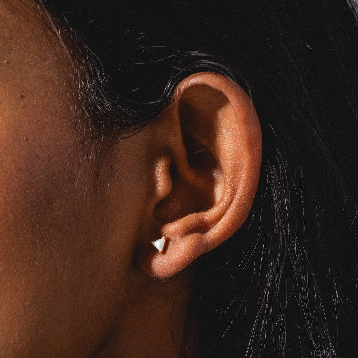 Close-up of a person’s ear with dark hair, wearing a small, triangular stud from the Classic Birthstone Earring Set. Lighting accentuates the skin texture and the earring’s metallic shine.