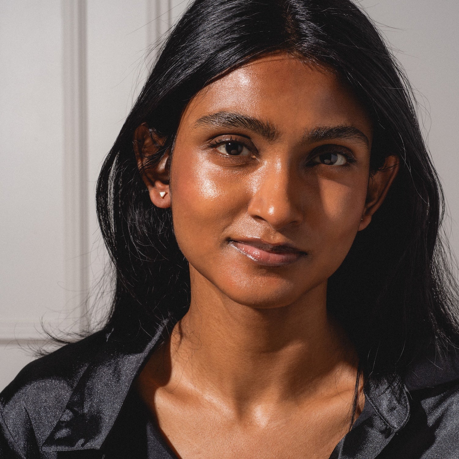 A woman with long black hair and brown skin wears a black top and the Classic Birthstone Earring Set, gazing gently at the camera as sunlight highlights her face against a light background.