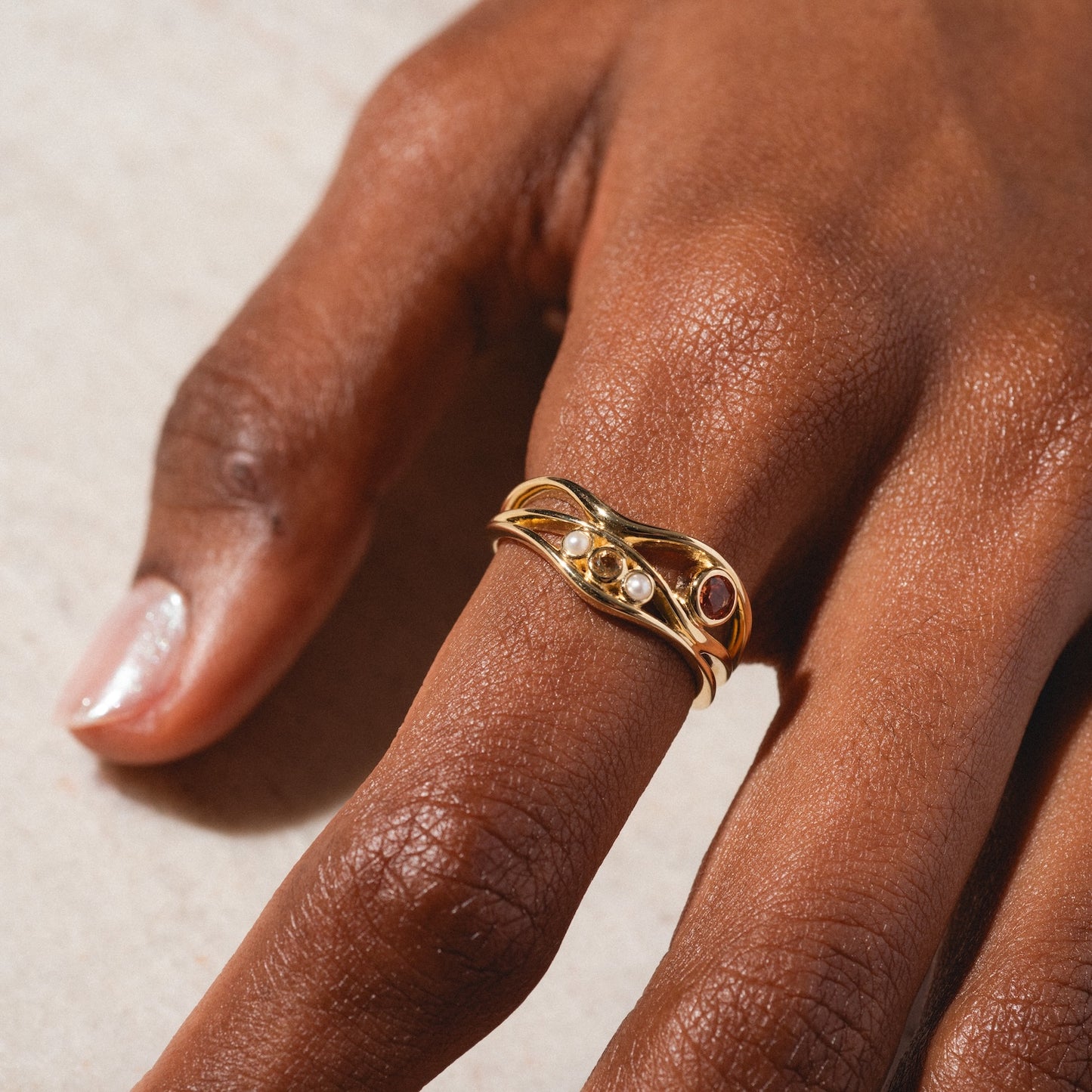 A close-up of a hand wearing the 10k Orange Sapphire and Citrine Bayside Ring, featuring intertwined gold bands with small white pearls and a single orange sapphire. The hand rests on a light surface, nails natural and unpolished.