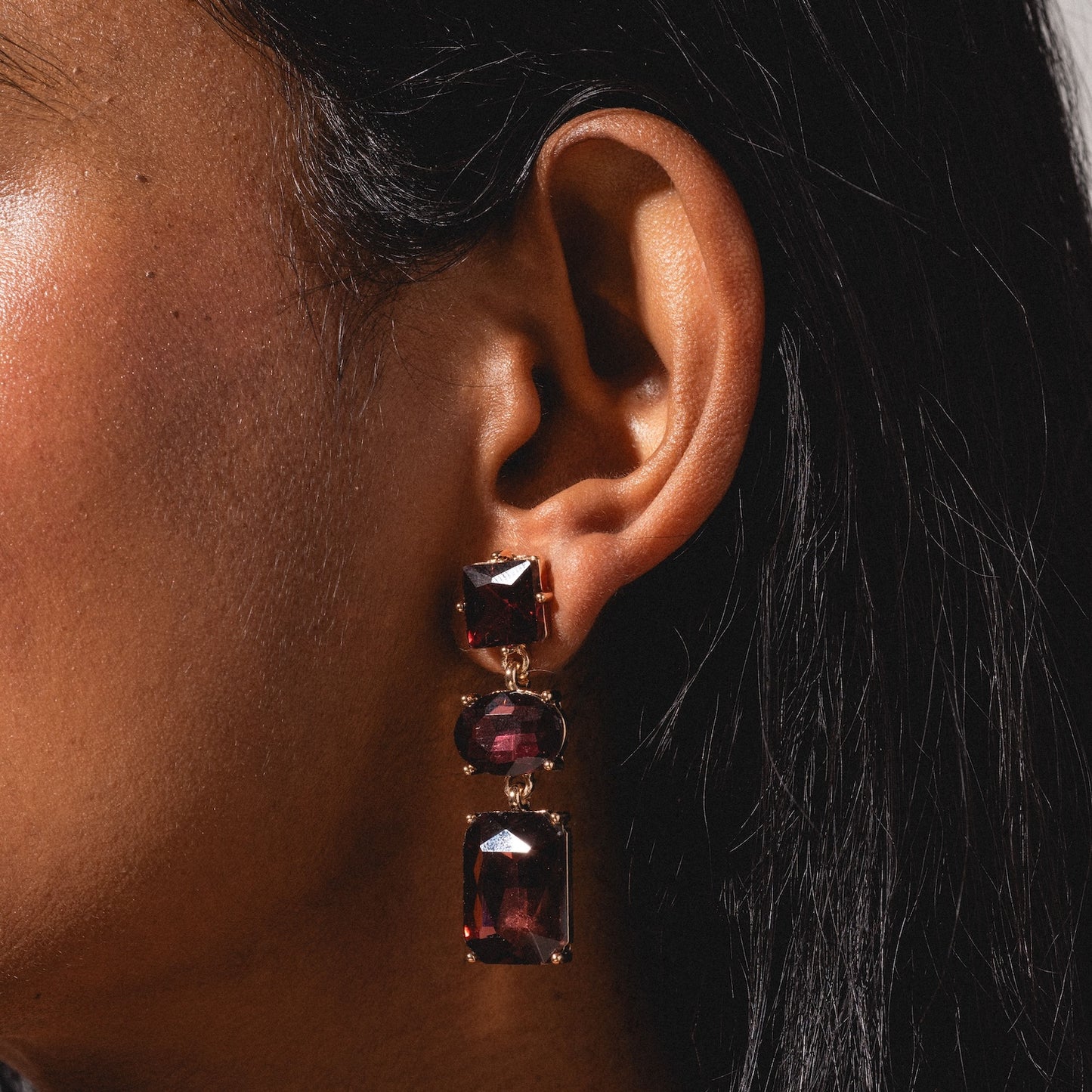 Close-up of a person’s ear with long, straight black hair, wearing the Allie Earrings—gold earrings featuring three large, uniquely shaped dark red gemstones.