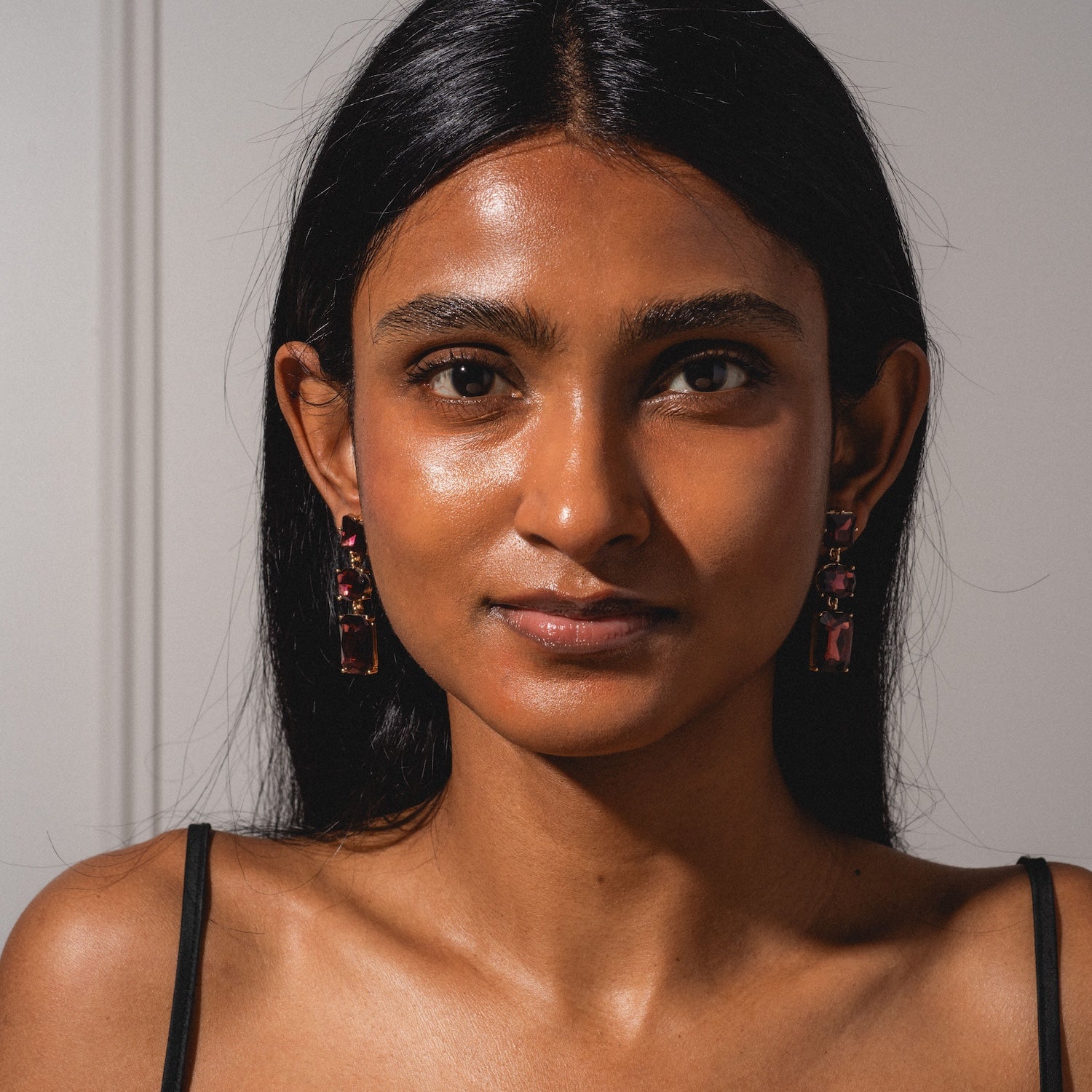 A woman with long dark hair and glowing skin faces the camera, wearing the bold red Allie Earrings and a black strap top, set against a neutral background.