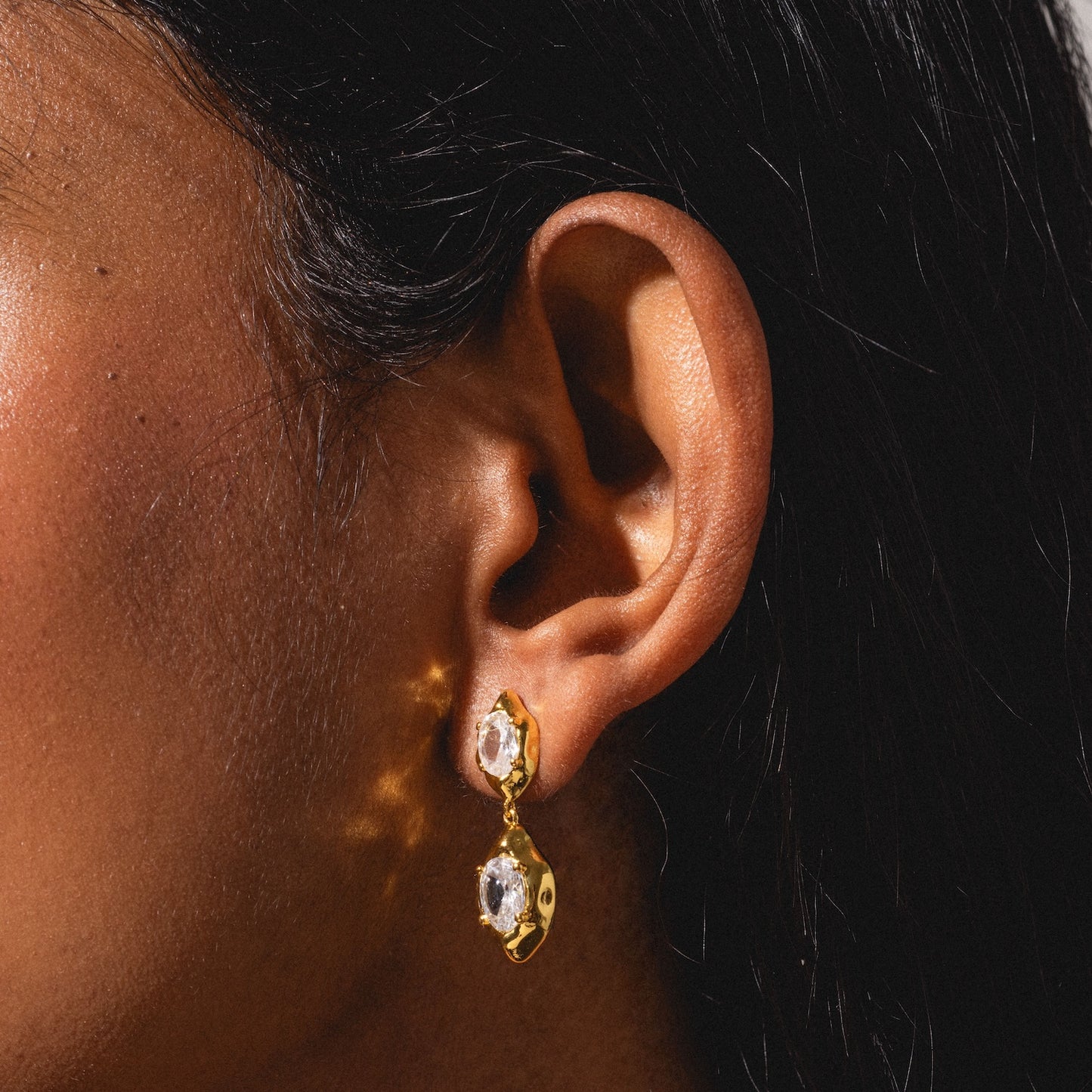 Close-up of a person’s ear wearing the Lowell Earrings—gold with two irregularly shaped, clear gemstones that reflect light onto the skin. The background is blurred.