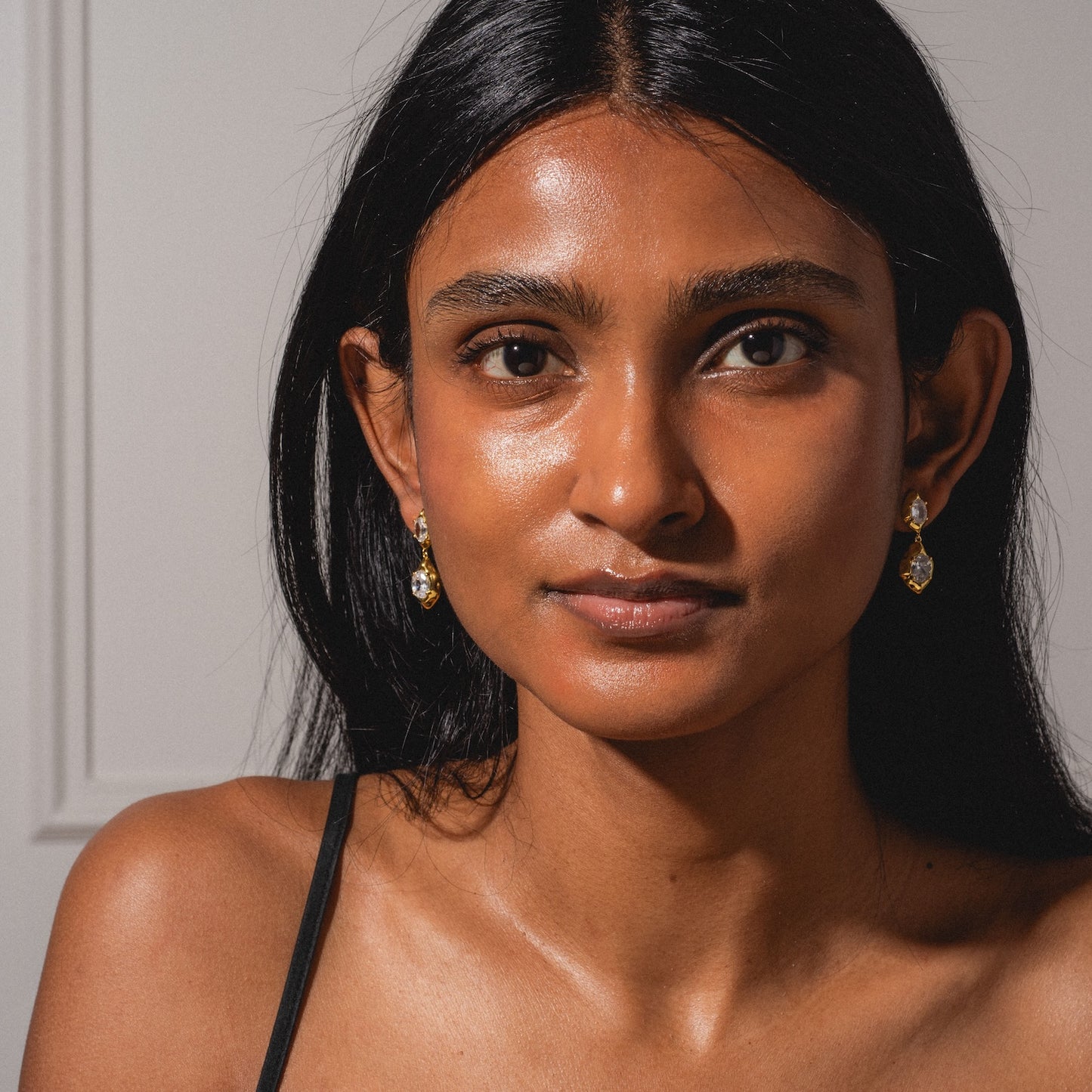 A woman with long black hair and glowing brown skin faces the camera, wearing the Lowell Earrings and a black strap top, set against a plain light background.