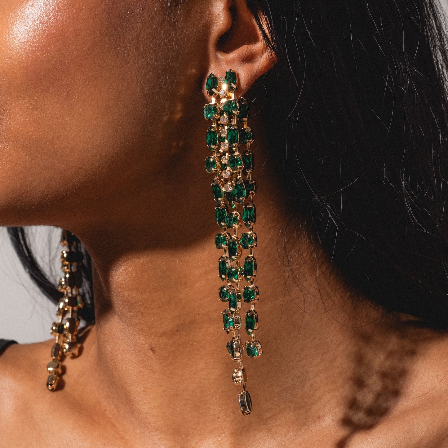 A close-up of a woman with straight dark hair wearing the Lola Earring—a long, gold piece adorned with multiple green gemstones—dangling by her neck. Only part of her face and shoulder are visible.