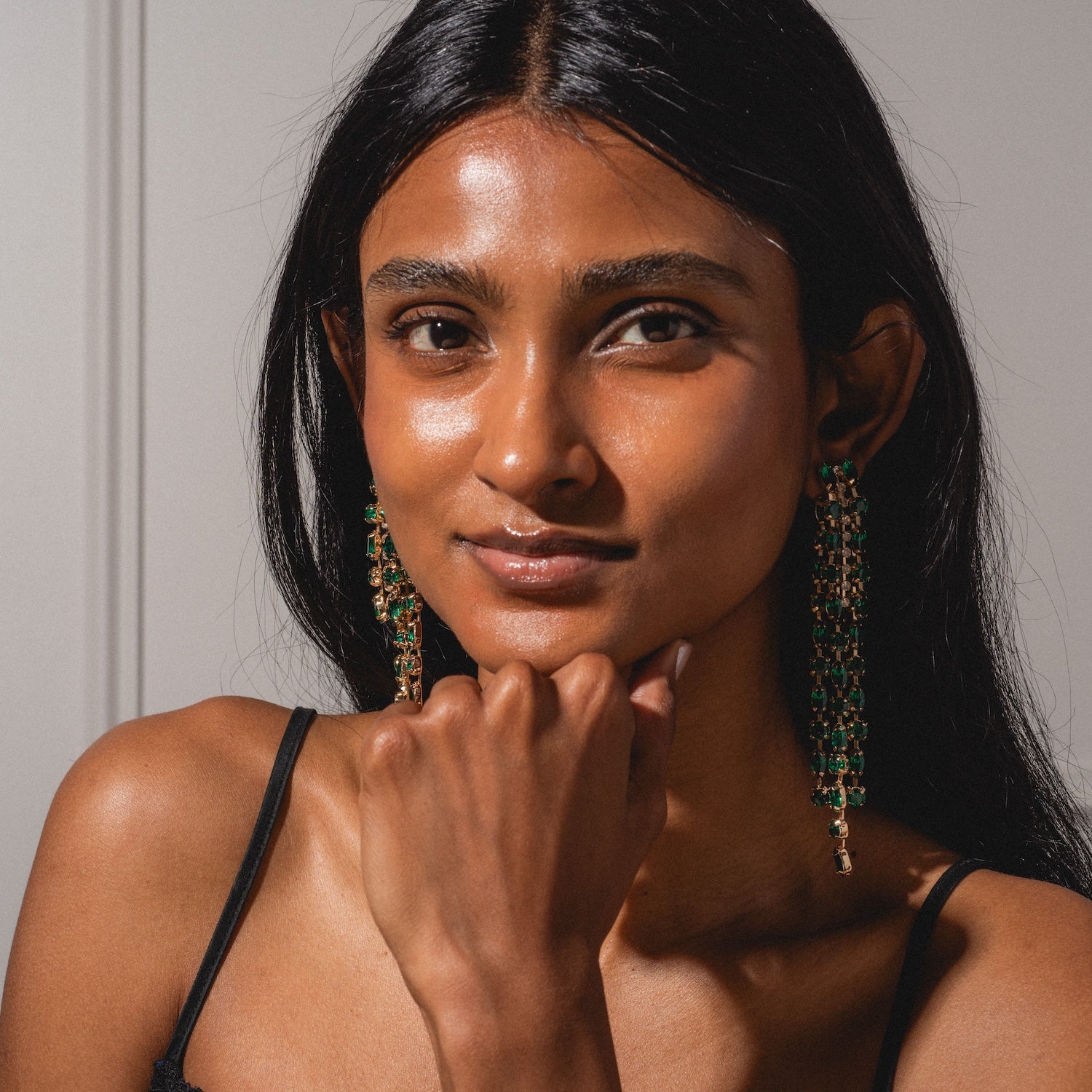 A woman with long dark hair and radiant skin wears the Lola Earring—dangling green and gold—paired with a black spaghetti-strap top, resting her chin on her hand and looking confidently at the camera.
