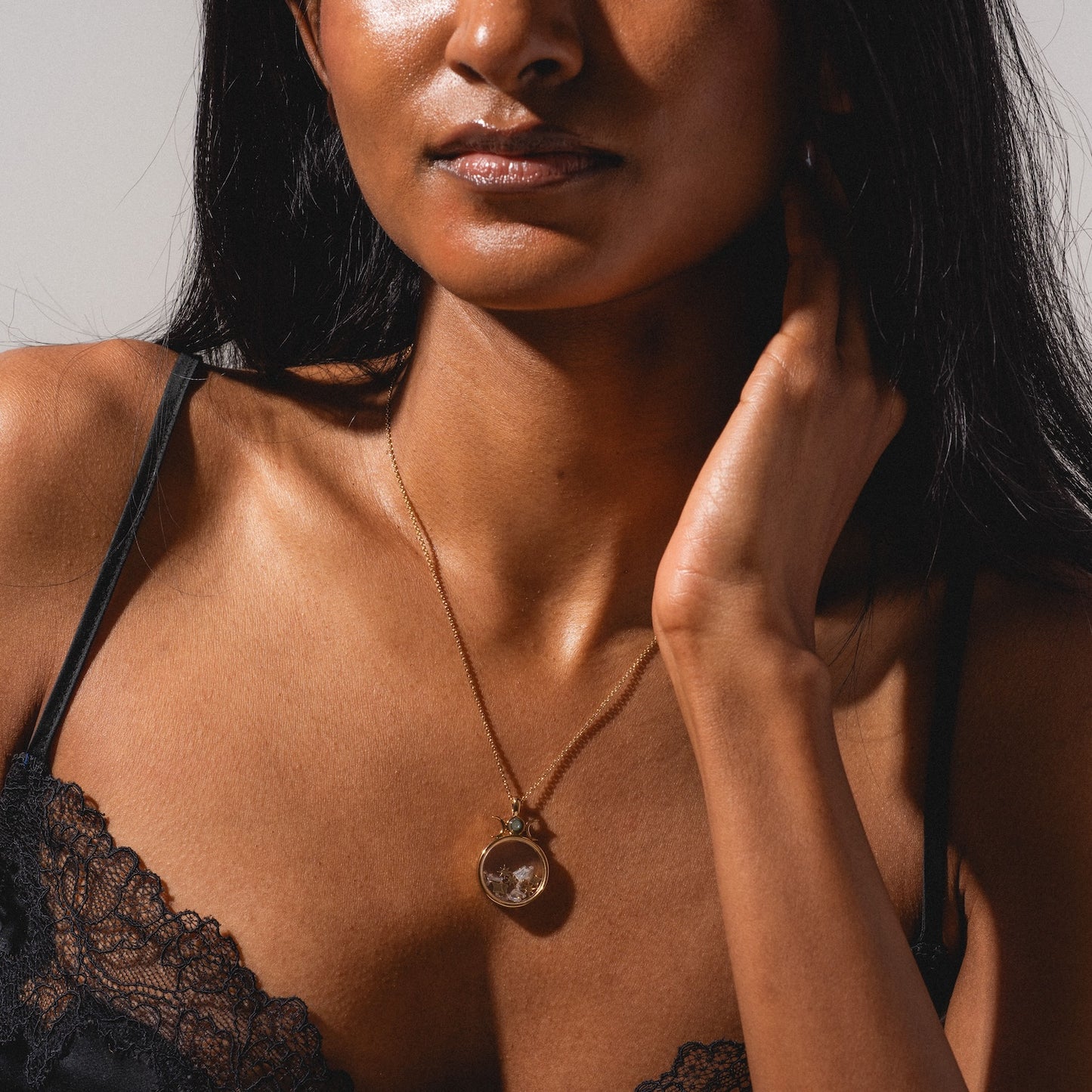 A woman in a black lace top gently touches her neck, showcasing the Wishkeeper Floating Locket Necklace. The image is cropped from shoulders to nose, highlighting the elegant jewelry and intricate lace details.