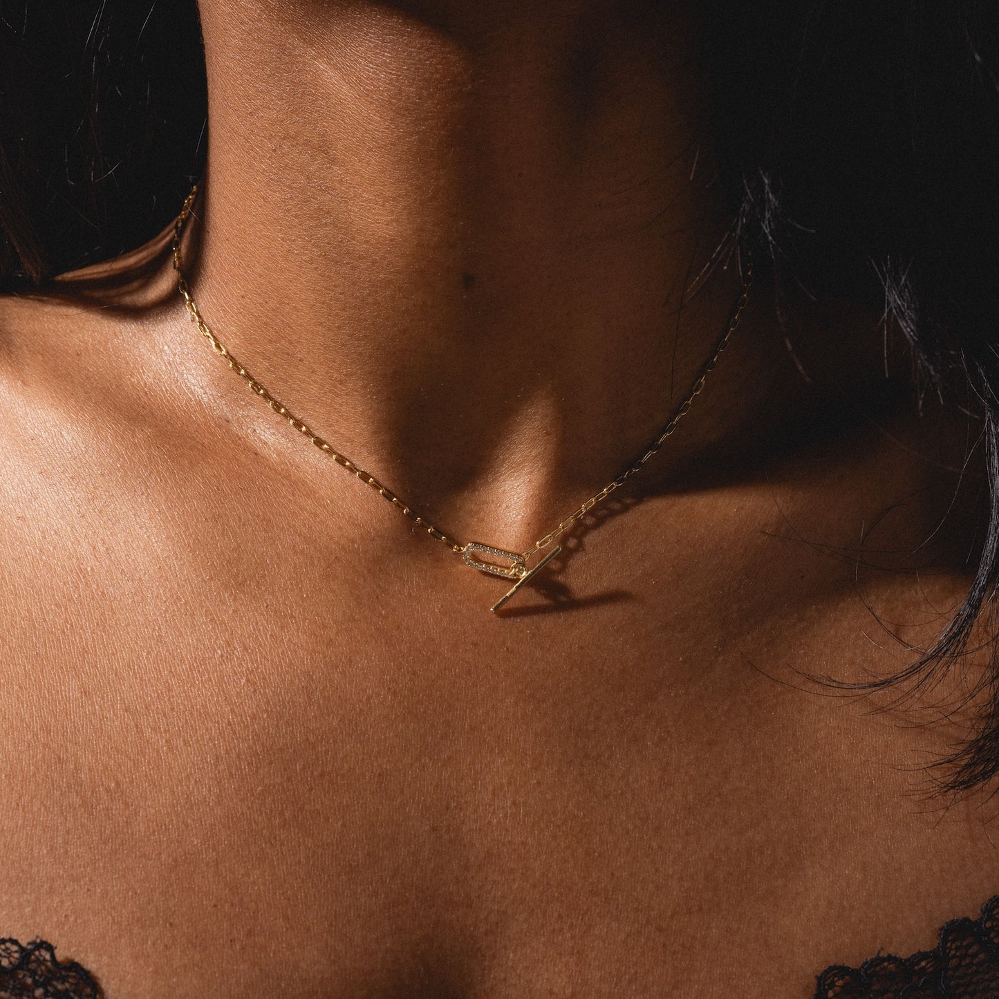 A close-up of a woman's neck wearing the Jade Pave Necklace—a delicate gold piece with a geometric pendant—gently lit by soft light. She is dressed in a black lace top.