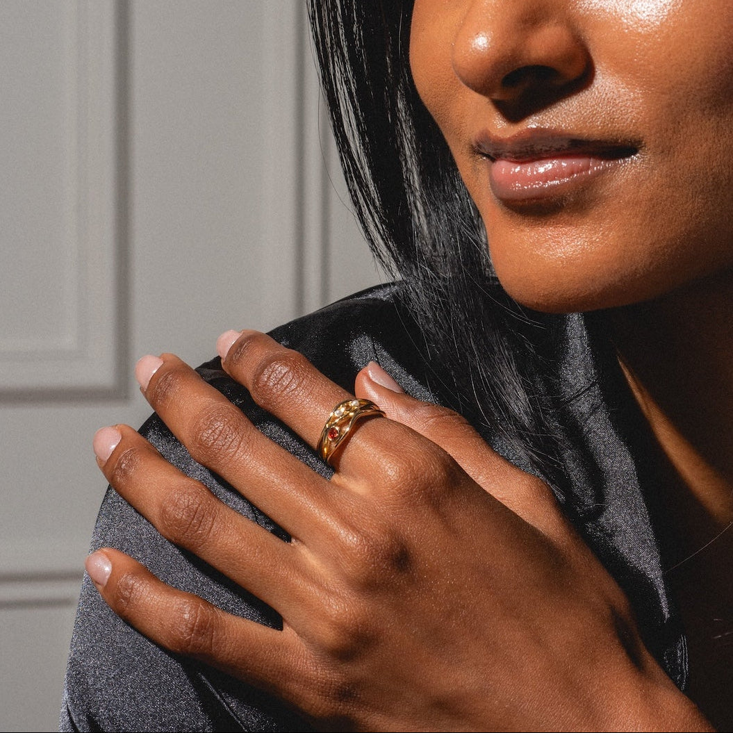 A woman with smooth, glowing skin and straight dark hair wears a dark shirt and displays the 10k Orange Sapphire and Citrine Bayside Ring on her hand, sunlight casting soft shadows as she looks at the camera.