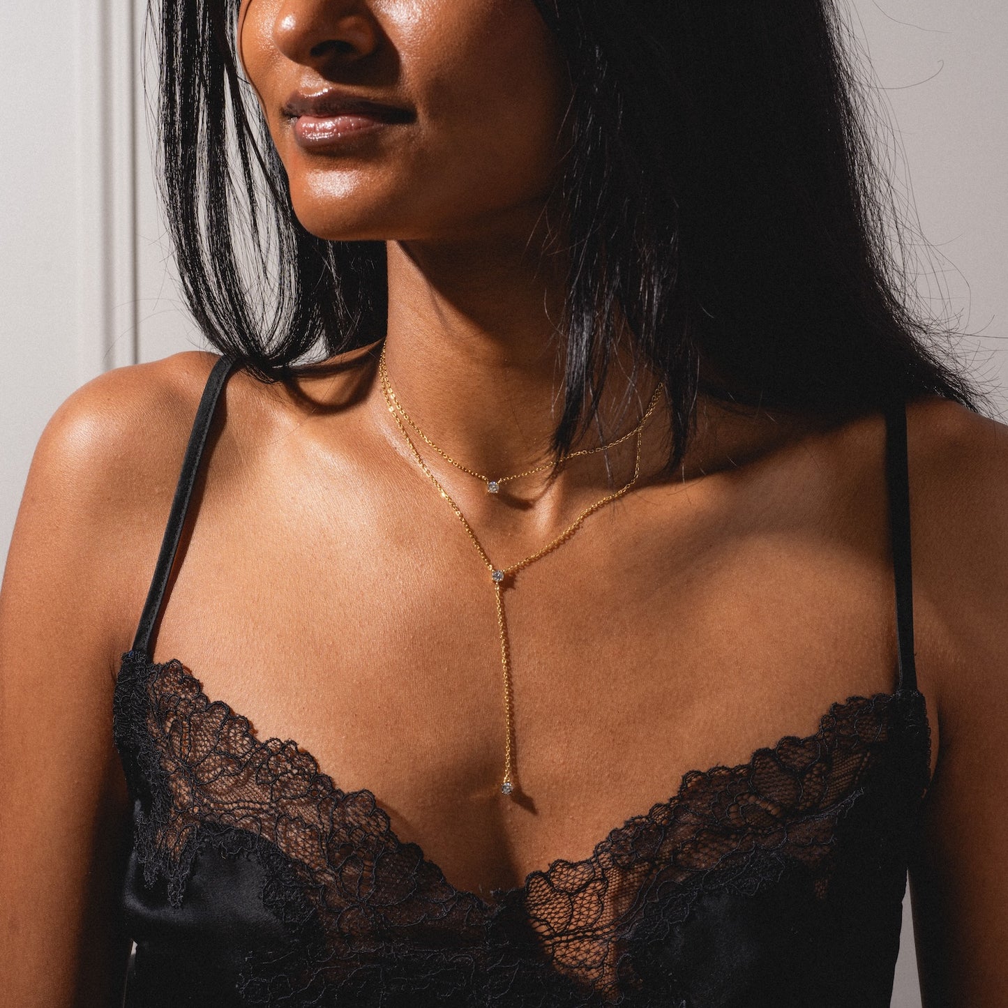 A woman wears the Solitaire Layered Lariat Necklace over a black lace camisole, posing against a neutral background to highlight the delicate gold layers and solitaire pendant.