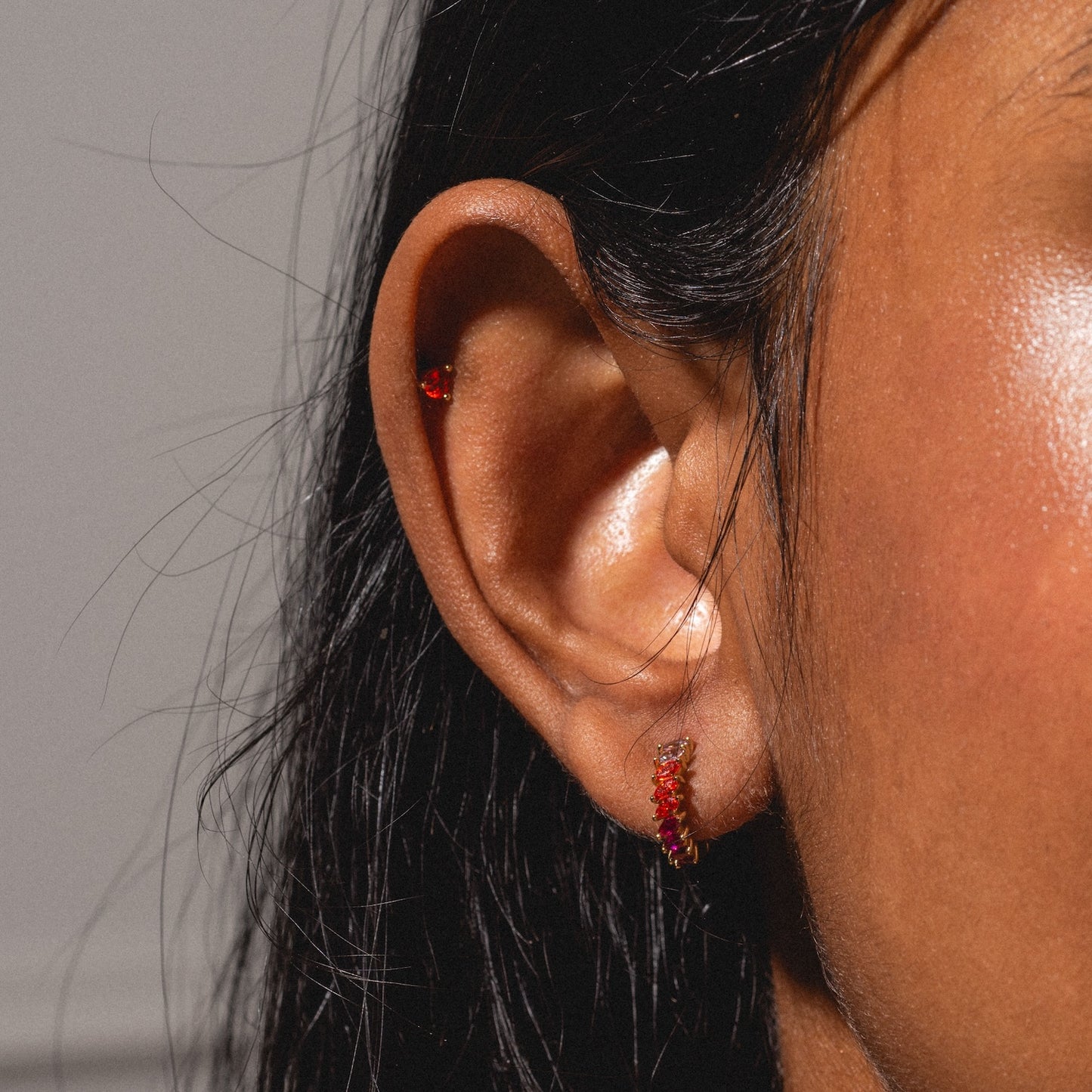 Close-up of a person with dark brown skin and straight black hair wearing the Bold Birthstone Earring Set: two gold earrings with small red stones, one in the earlobe and one in the cartilage.