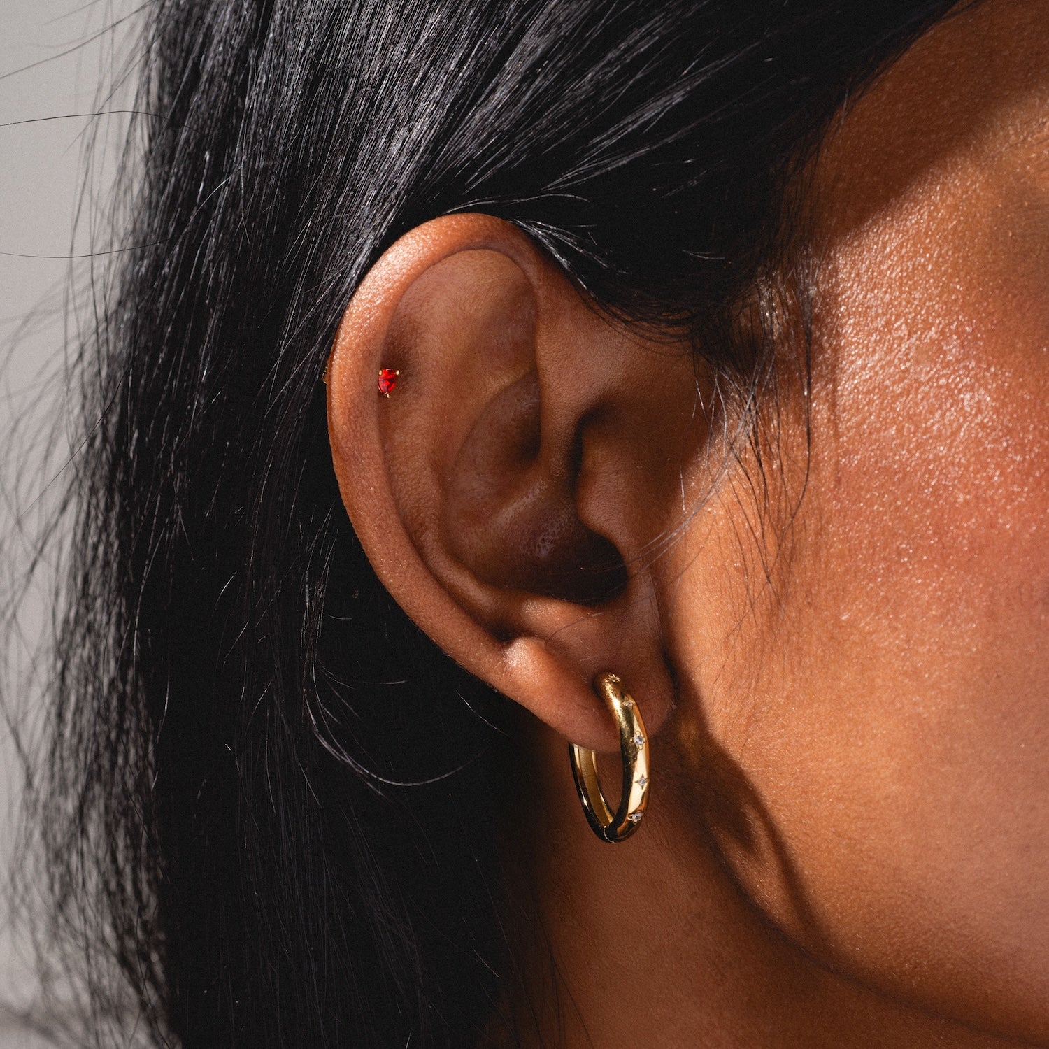 A close-up of a person's ear wearing the Bold Birthstone Earring Set: a small red stud in the upper cartilage and a gold hoop in the lobe. They have straight black hair and medium brown skin.