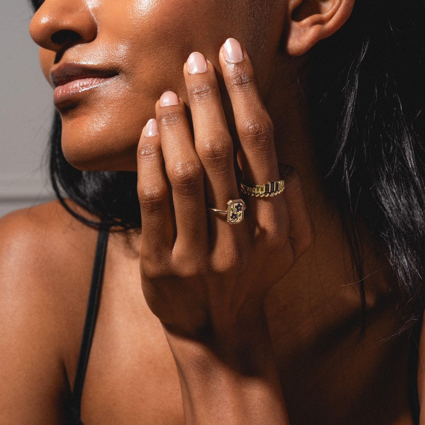 A woman with radiant skin touches her face, showcasing neatly manicured nails and two gold rings, including the 10k Hidden Message Morse Code Diamond Band. She wears a black strap; soft lighting highlights her jewelry.