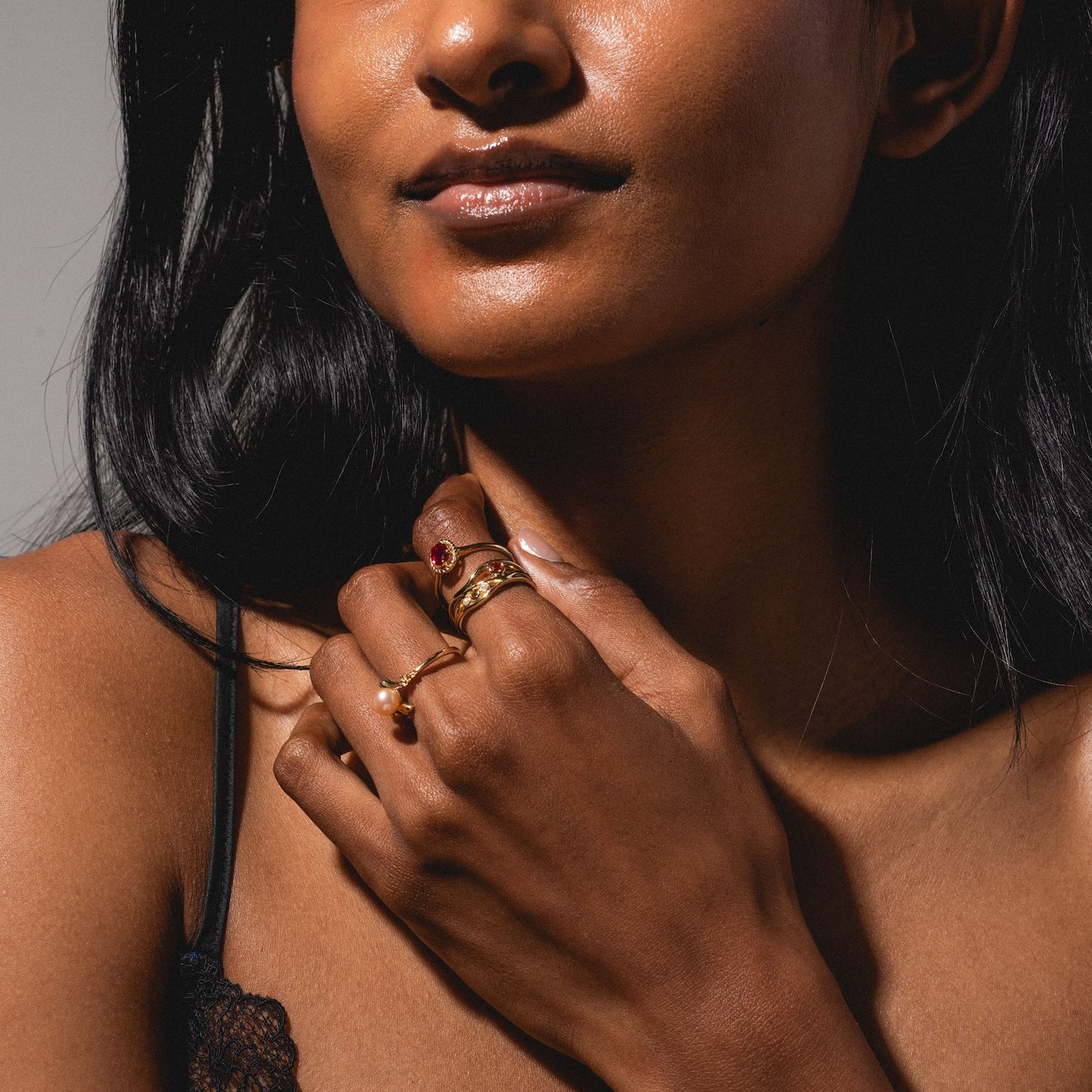 A woman with long dark hair wears a black lace strap top and several gold rings, including the 10k Lucky In Red Ruby Halo Ring, as she poses with her hand near her chin and her lips looking slightly glossy.