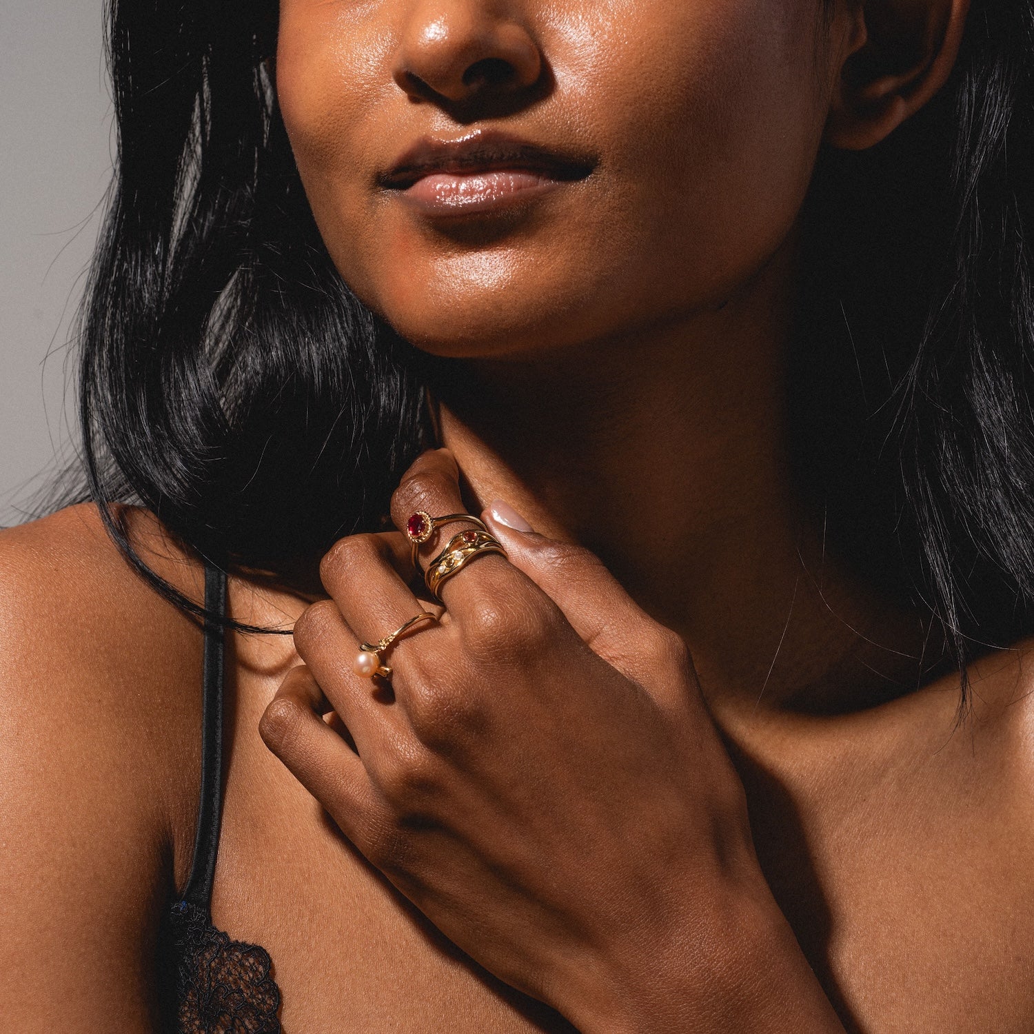 A woman with long dark hair wears a black lace strap top and several gold rings, including the 10k Lucky In Red Ruby Halo Ring, as she poses with her hand near her chin and her lips looking slightly glossy.