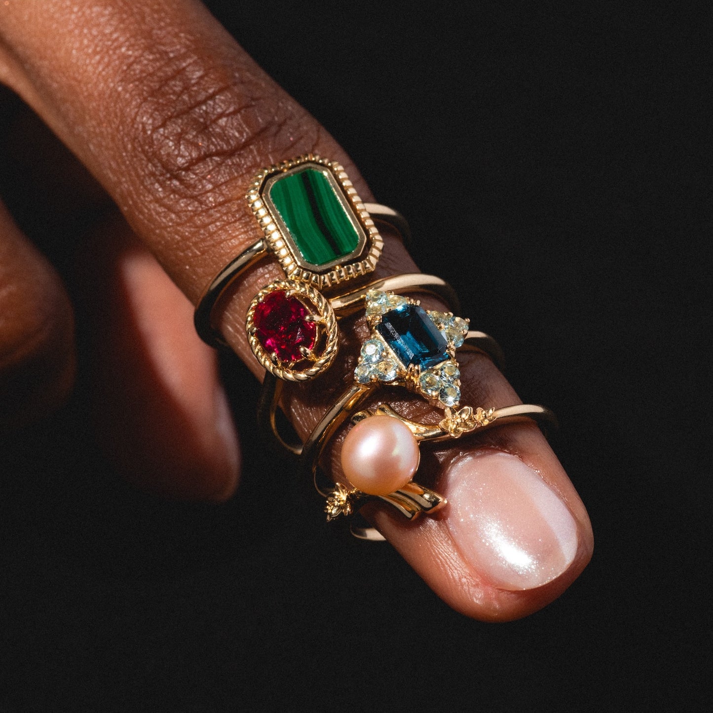 A hand wears the 10k Gallery Frames Enamel and Malachite Reversible Ring—one of four gold rings with distinct gemstones (green, red, blue with diamonds, and a pink pearl)—all stacked on one finger against a black background.