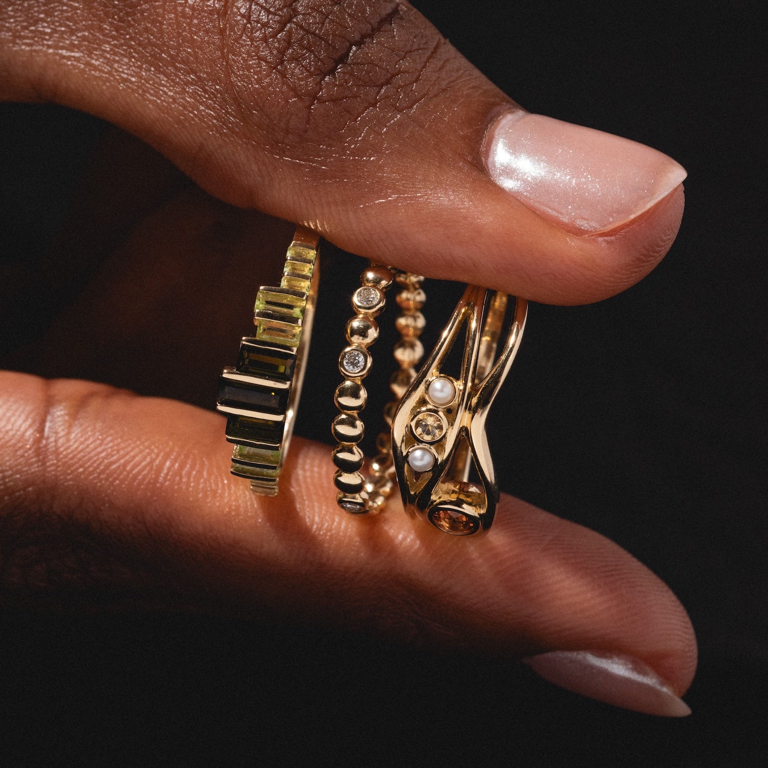 A hand with a light pink manicure holds three ornate gold rings, including the 10k Hidden Message Morse Code Diamond Band, each with unique gemstones and designs, set against a dark background.