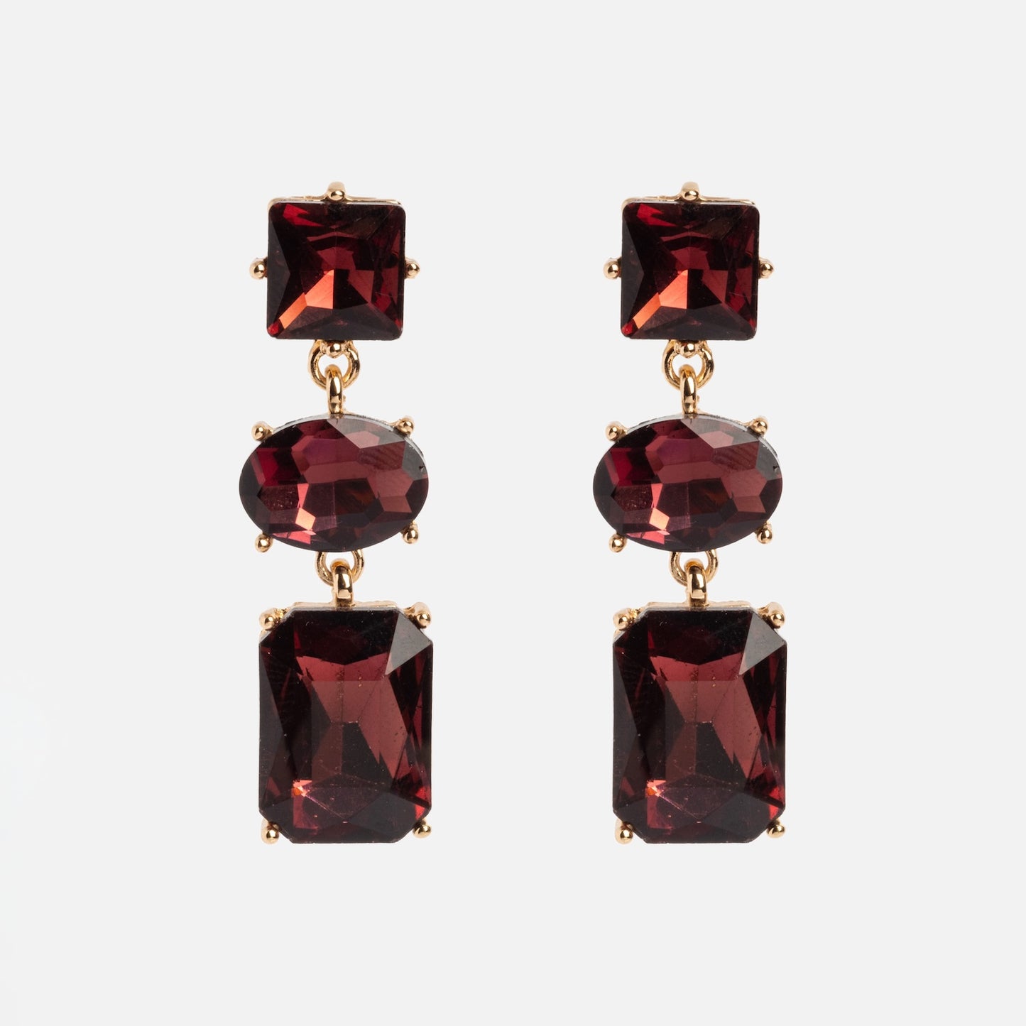 The Allie Earrings are gold drop earrings featuring three faceted red gemstones—square, oval, and rectangular—set vertically against a white background.