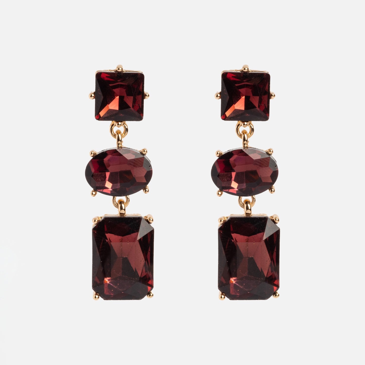 The Allie Earrings are gold drop earrings featuring three faceted red gemstones—square, oval, and rectangular—set vertically against a white background.