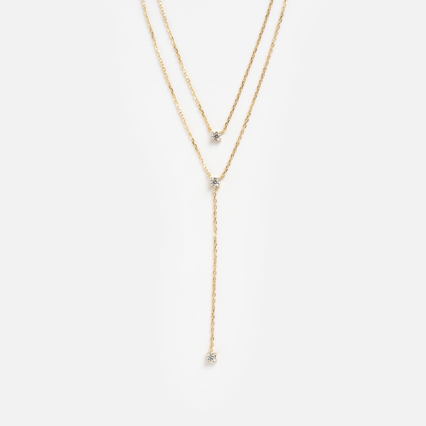 The Solitaire Layered Lariat Necklace features two fine gold chains: one sitting higher, both accented with a single gemstone, and the longer chain finishing in a vertical lariat style with a gemstone drop.