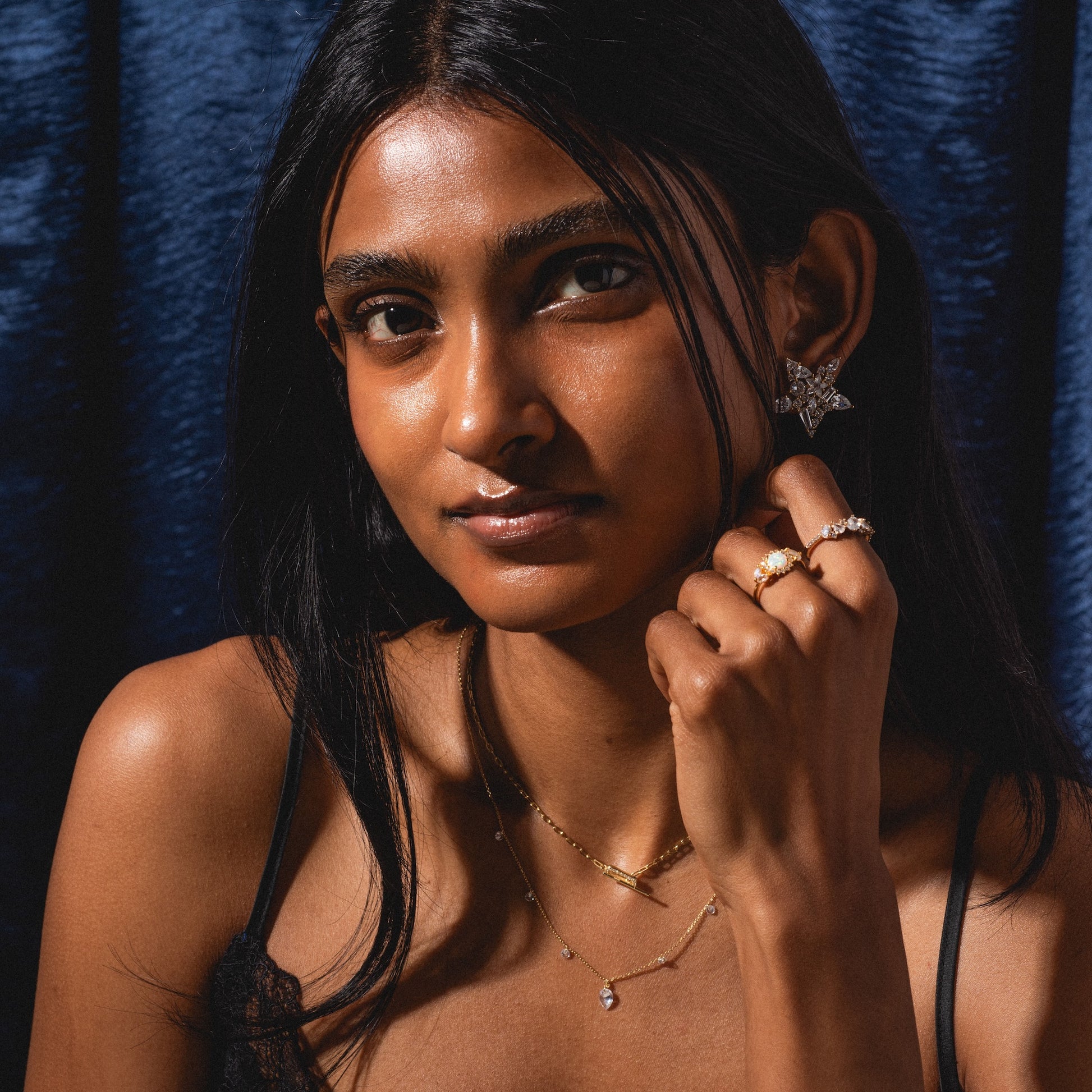 A woman with long dark hair poses before a dark blue background, wearing a black top and the Floating CZ Station Necklace, styled with multiple rings and star-shaped earrings.