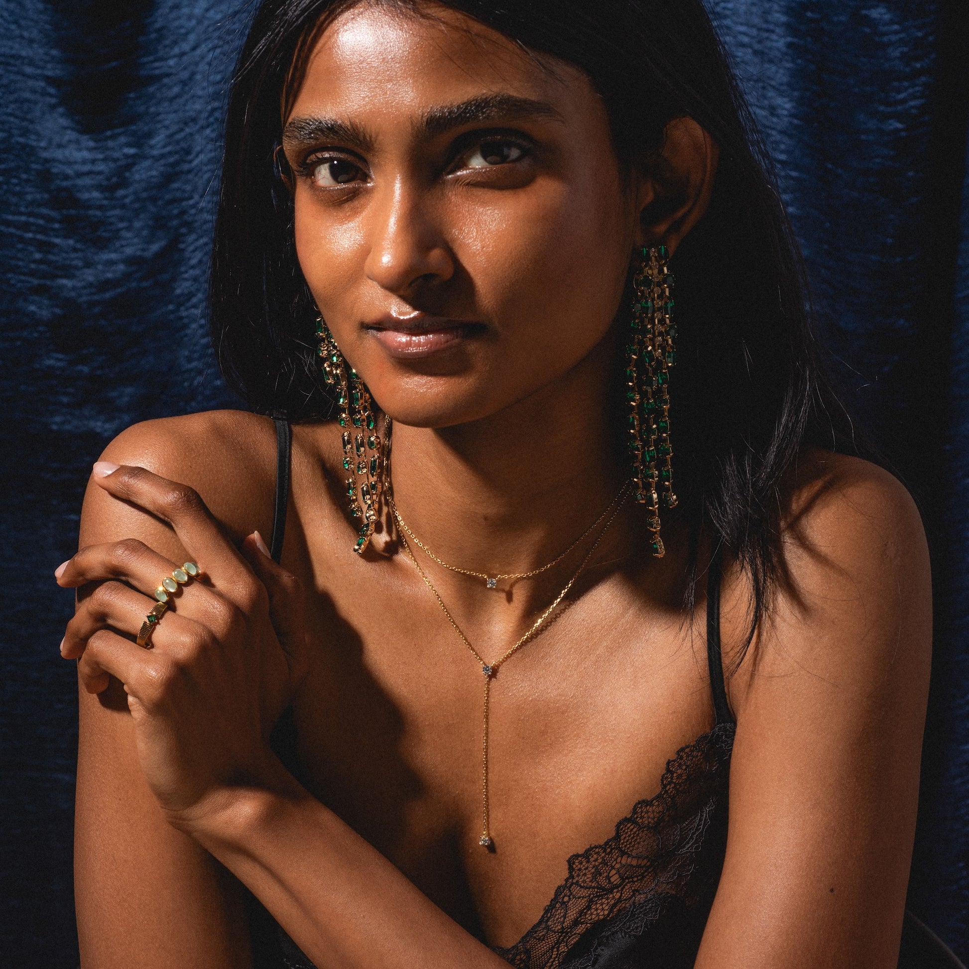 A woman with long dark hair poses against a dark blue background. She wears a black lace top, ornate green and gold earrings, gold rings, and the Solitaire Layered Lariat Necklace, with her arms gently crossed.