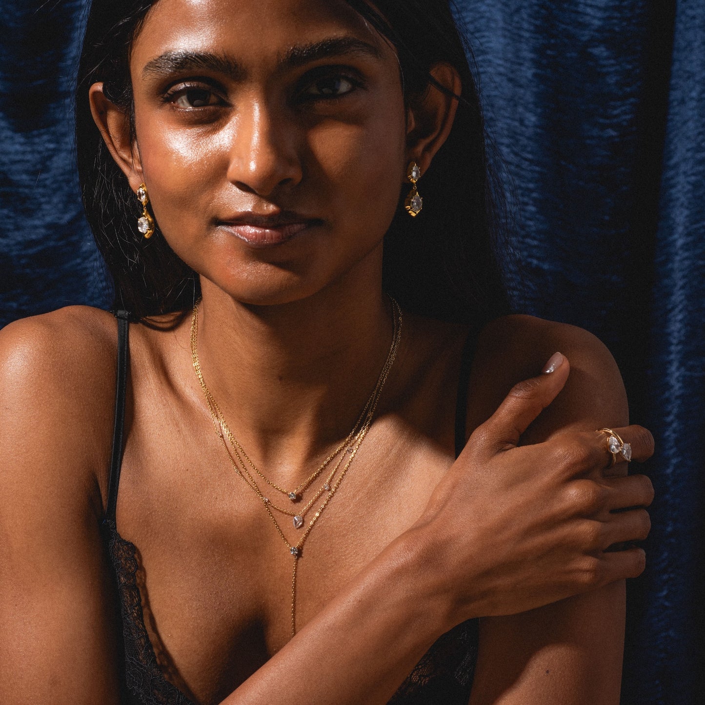 A woman with long dark hair, dressed in a black lace top and gold drop earrings, poses against a dark blue velvet background. She wears the Solitaire Layered Lariat Necklace and gently rests her hand on her shoulder.