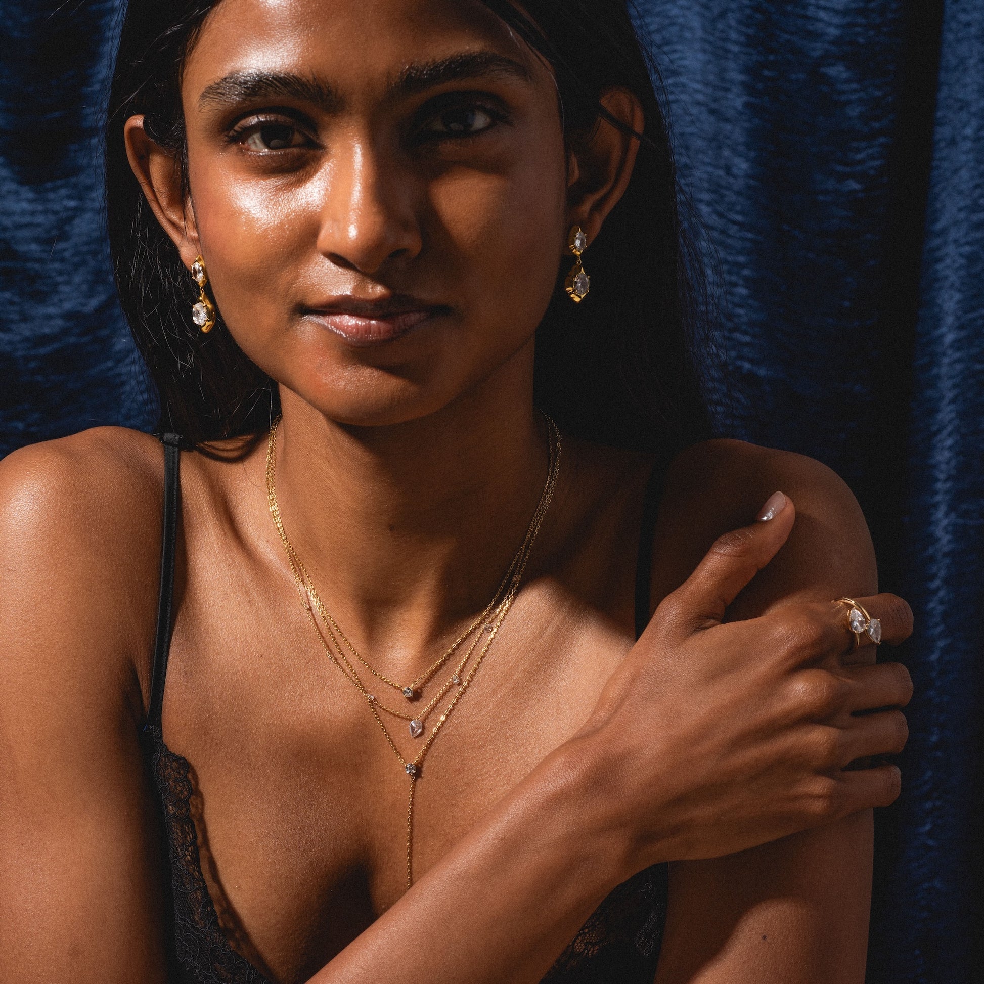 A woman with long dark hair, dressed in a black lace top and gold drop earrings, poses against a dark blue velvet background. She wears the Solitaire Layered Lariat Necklace and gently rests her hand on her shoulder.