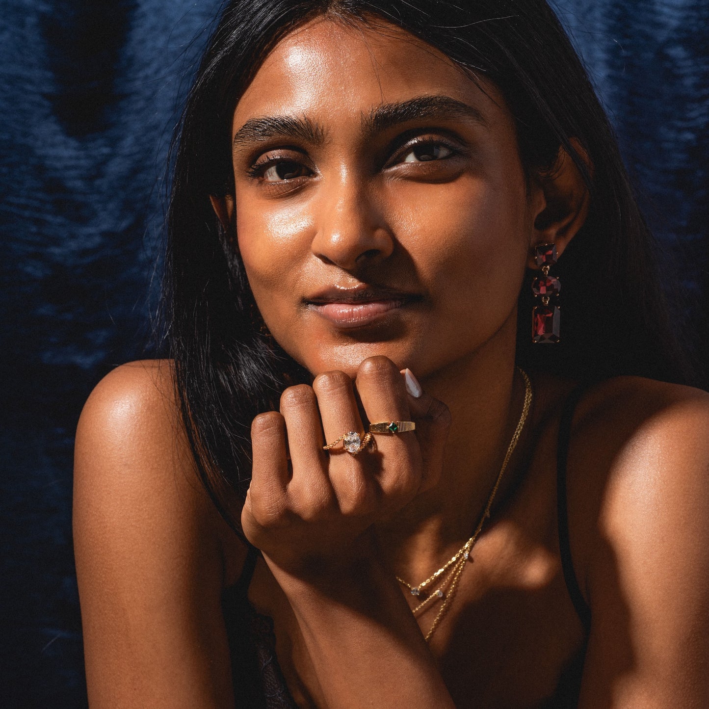 A woman with long dark hair poses against a dark blue background, resting her chin on her hand. She wears gold necklaces, rings, and the bold Allie Earrings, with natural makeup and a calm expression.