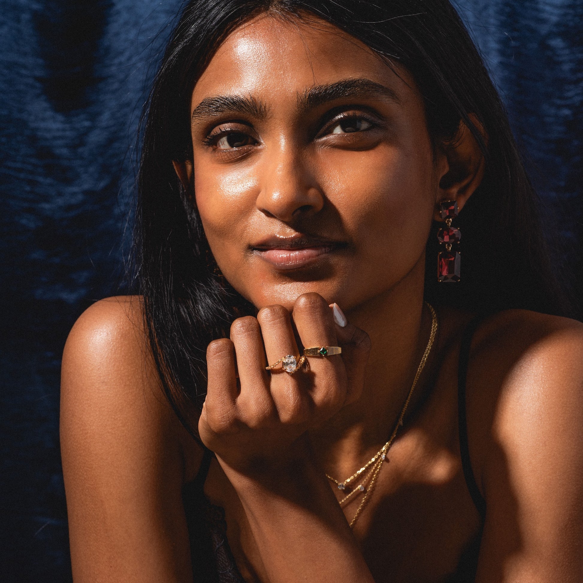 A woman with long dark hair poses against a dark blue background, resting her chin on her hand. She wears gold necklaces, rings, and the bold Allie Earrings, with natural makeup and a calm expression.
