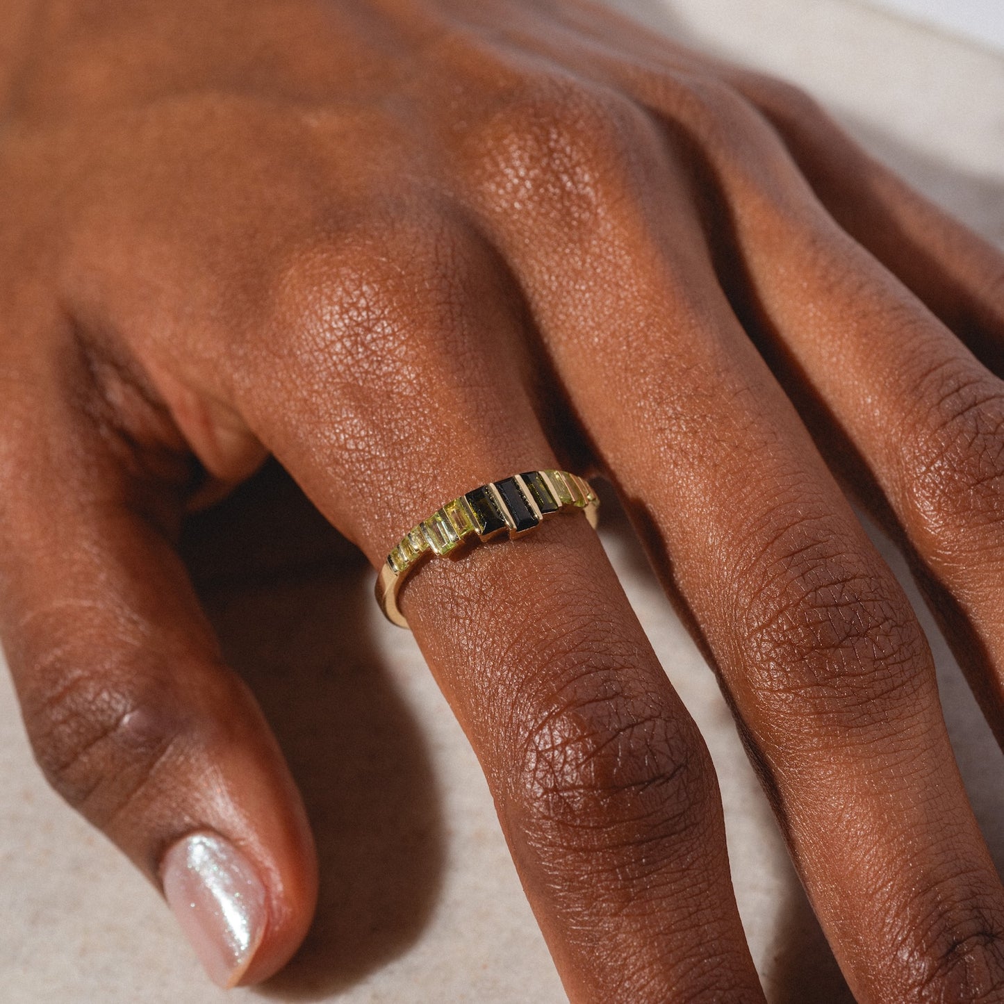 A close-up of a hand with brown skin wearing the 10k Green Tourmaline Skyline Band—its gold design features vertical rectangular segments. The nails are neatly manicured, with one painted in a shimmery light shade.