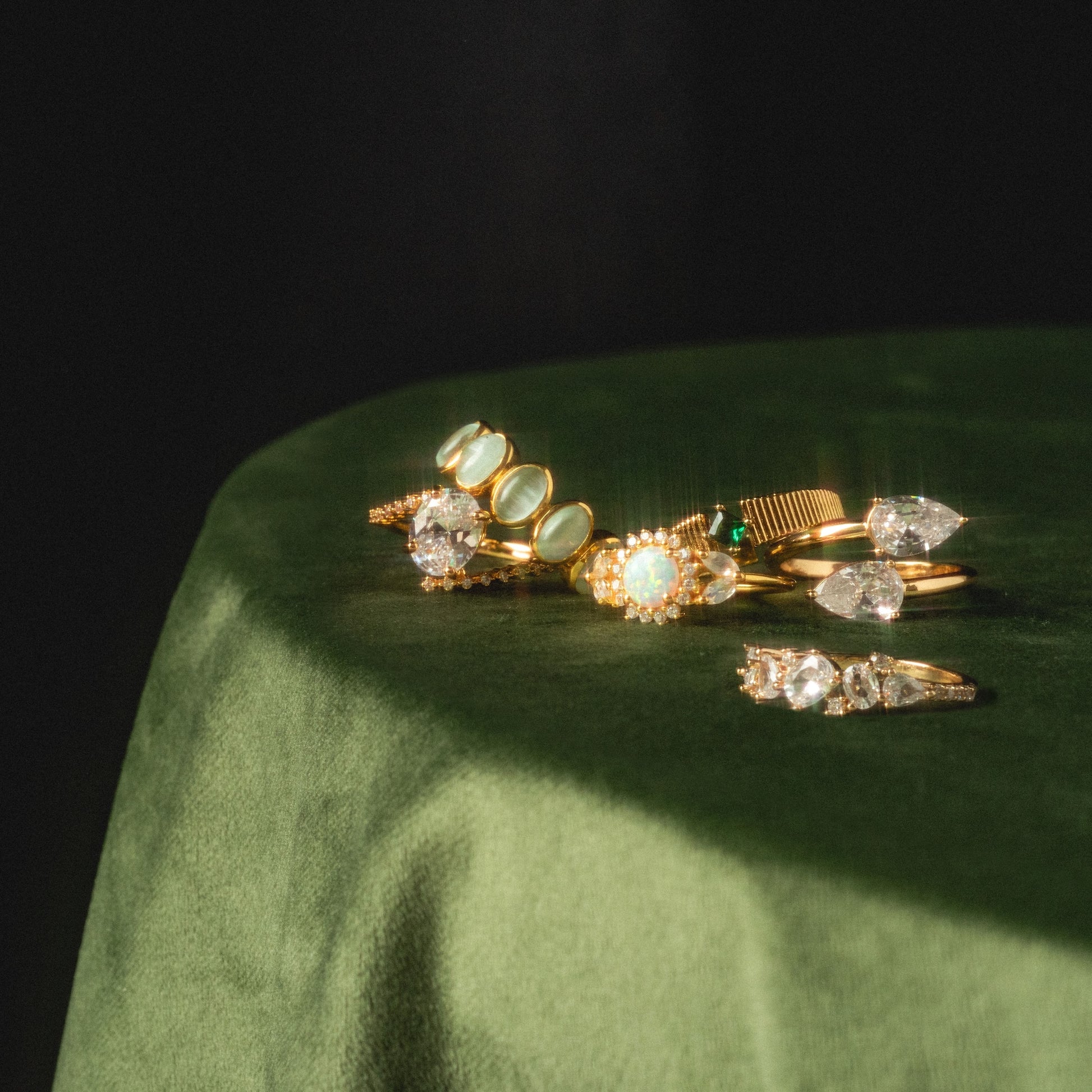 The Tilted CZ Ring is displayed with other gold rings featuring various gemstones and diamond settings on a round, green velvet surface in soft, natural light.
