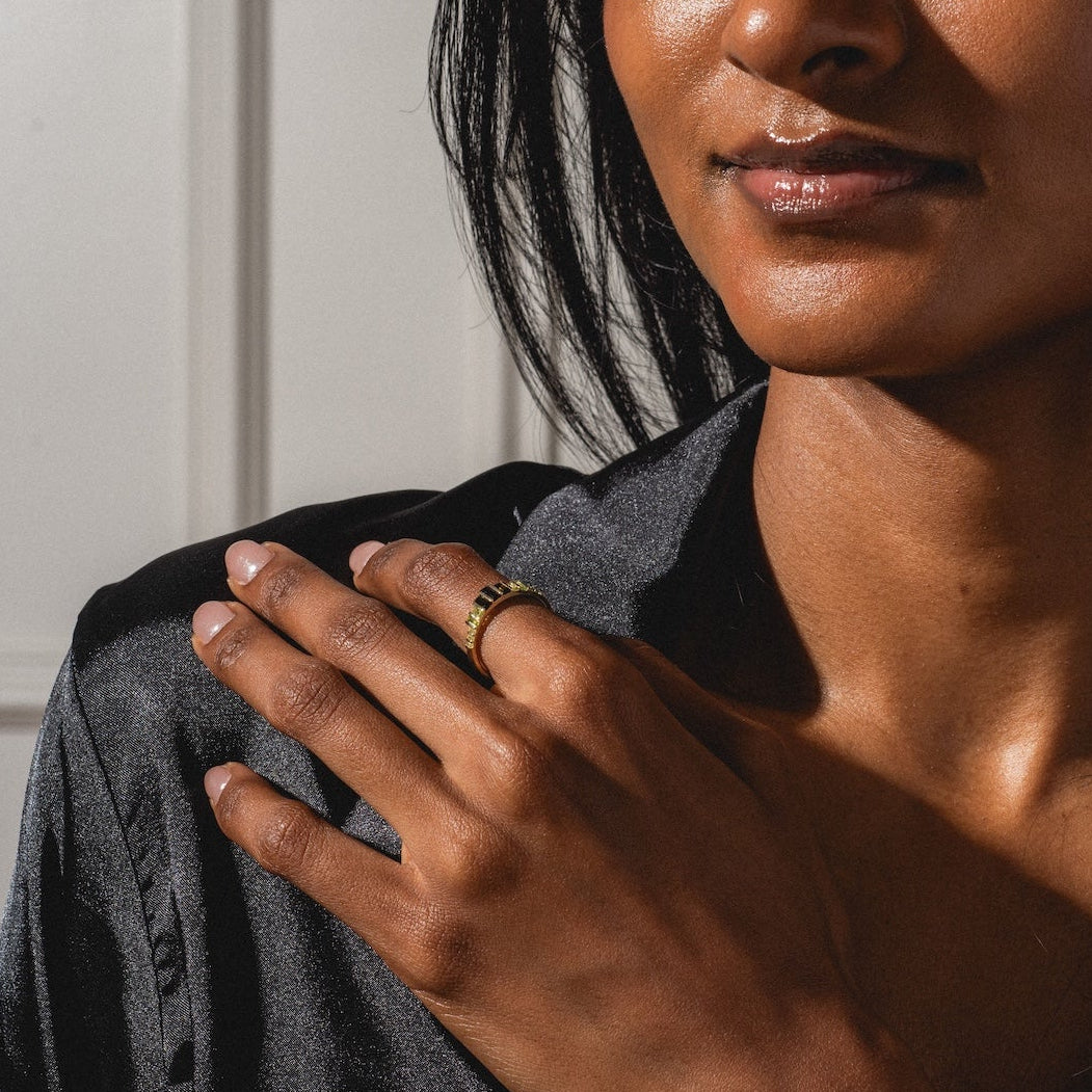 A woman with long dark hair in a black shirt poses indoors, resting her hand on her shoulder and confidently displaying the 10k Green Tourmaline Skyline Band as sunlight highlights her face and hand.